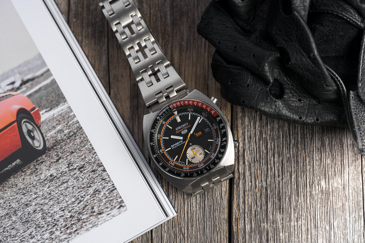 Seiko Speedtimer chronograph watch with stainless steel bracelet, black dial, orange accents, and tachymeter bezel on wooden surface