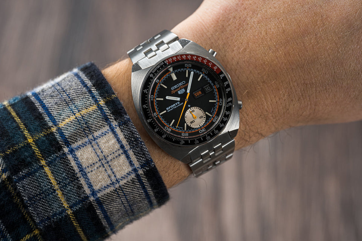 Seiko Speedtimer chronograph watch with black dial, orange accents, and stainless steel bracelet on wrist with plaid shirt