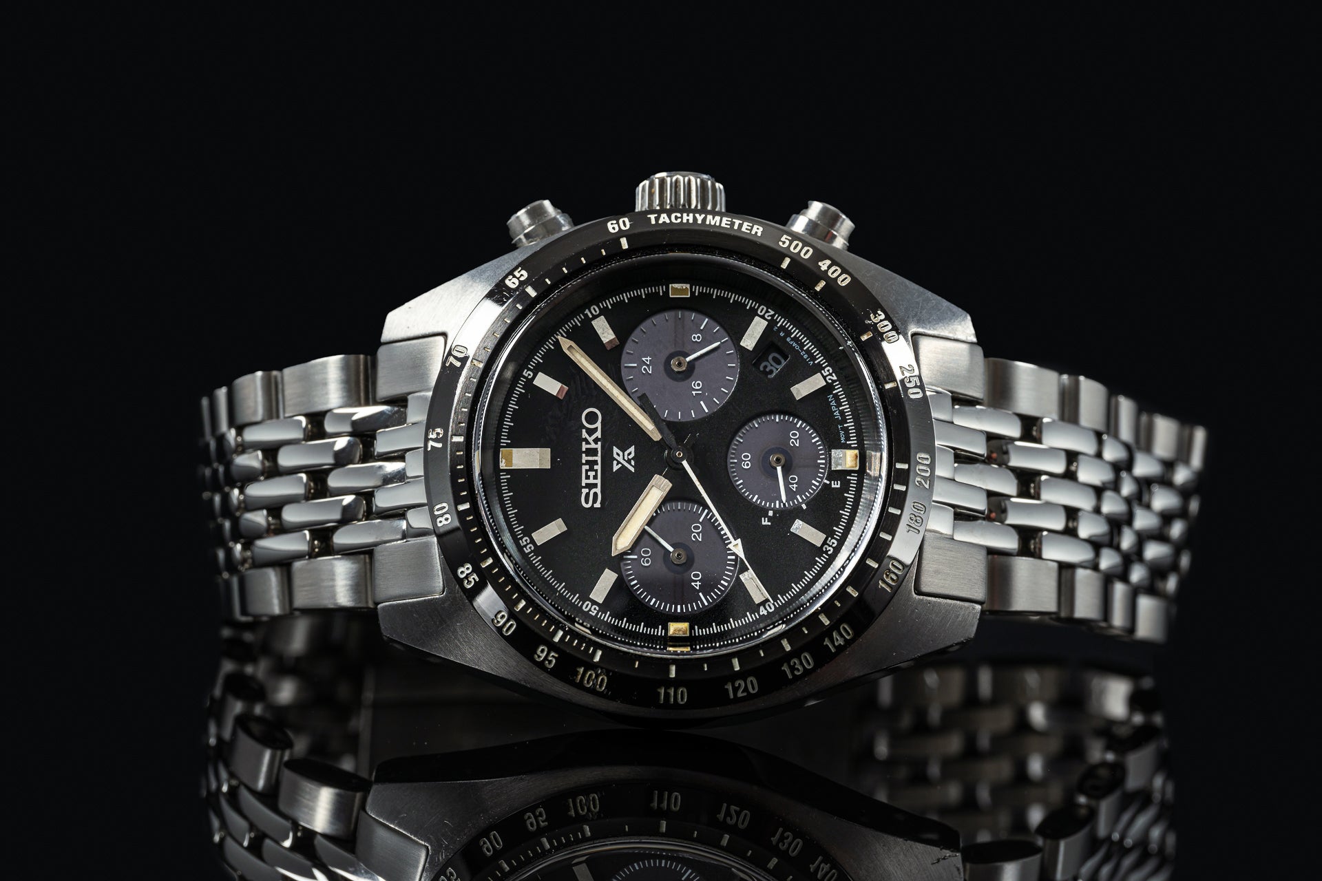 Seiko chronograph watch with black dial, tachymeter bezel, three subdials, and stainless steel beads of rice bracelet on black background