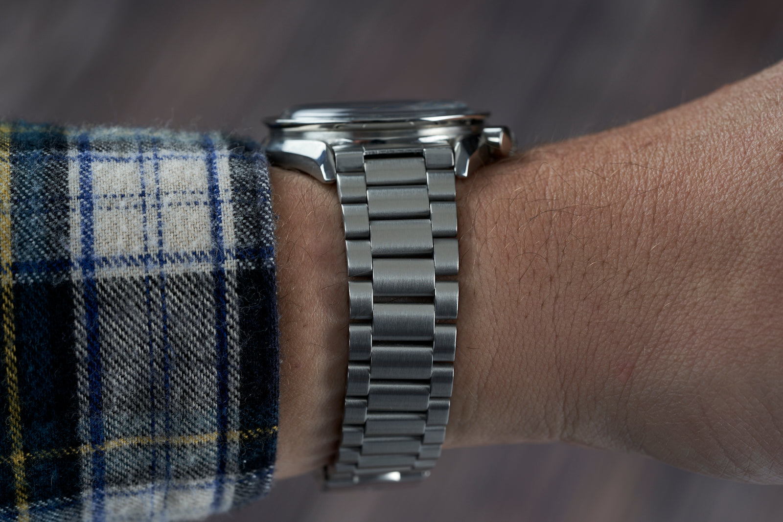 Stainless steel bracelet with polished links worn on wrist with plaid shirt sleeve, professional watch strap detail