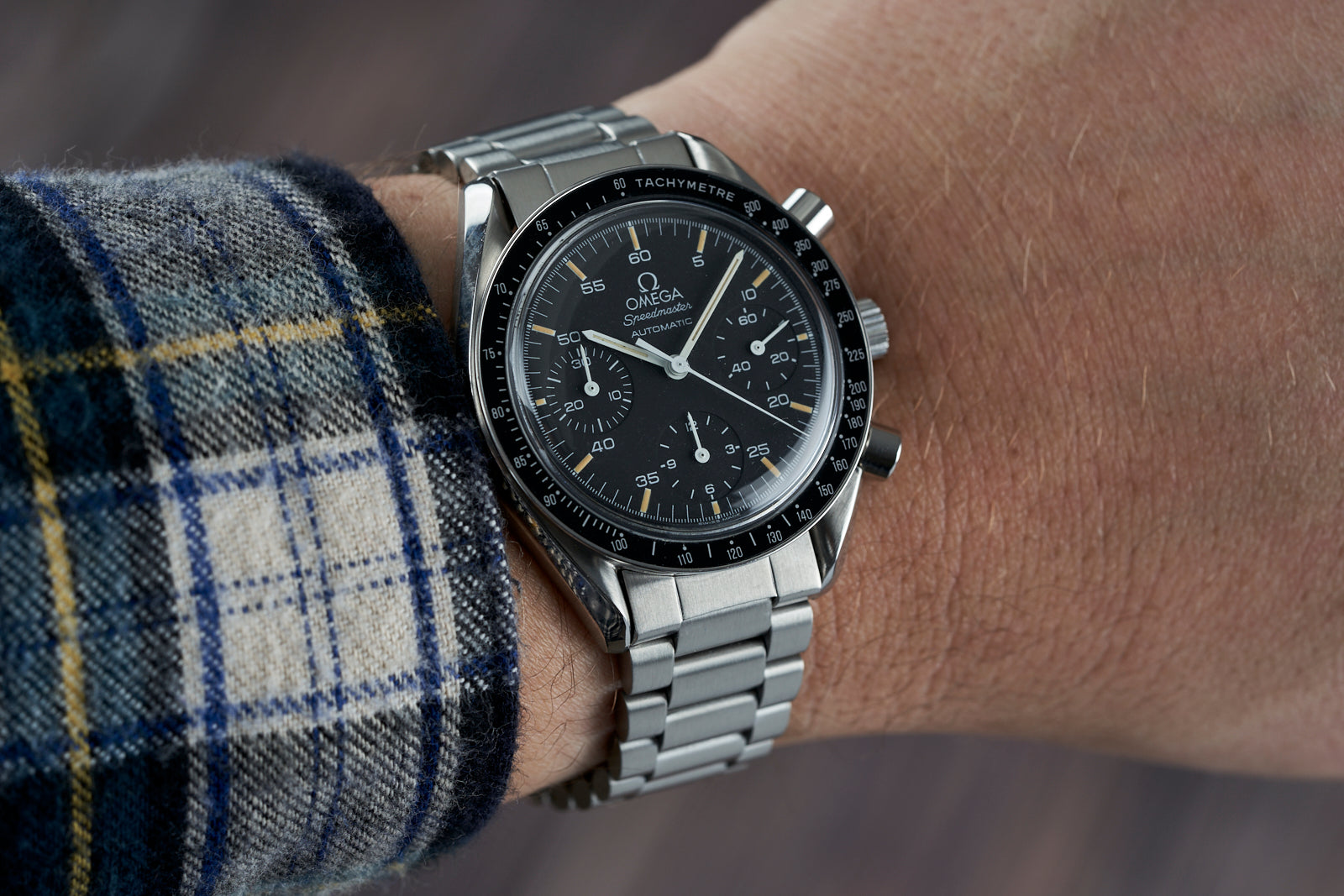 Omega Speedmaster Reduced chronograph watch with stainless steel bracelet and black dial on wrist, wearing plaid sleeve