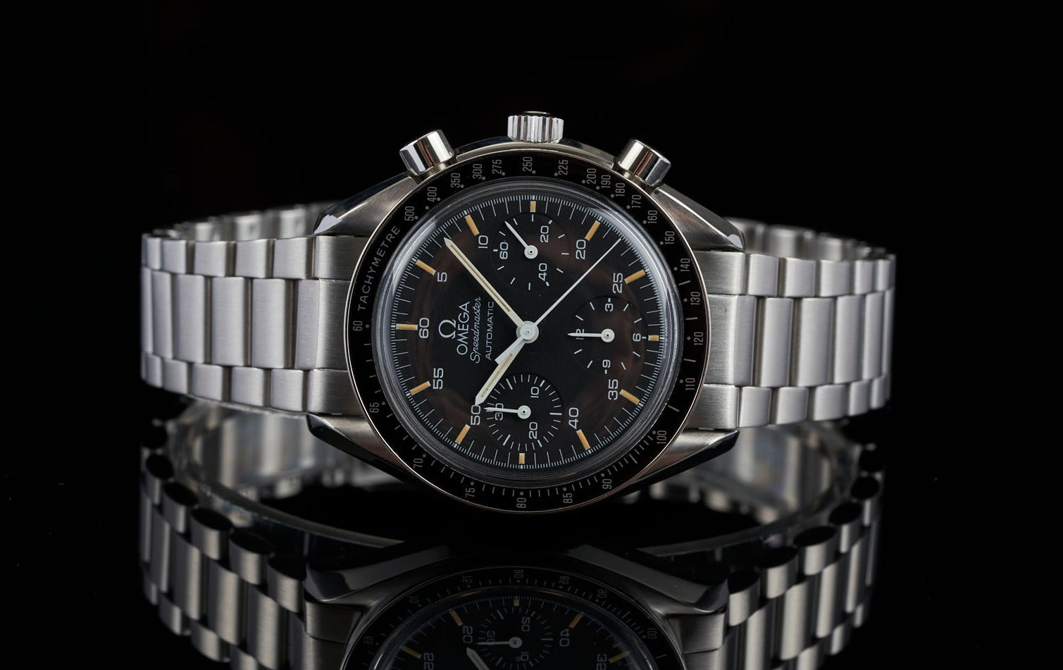 Omega Speedmaster Reduced chronograph watch with black dial, steel bracelet and tachymeter bezel on reflective surface