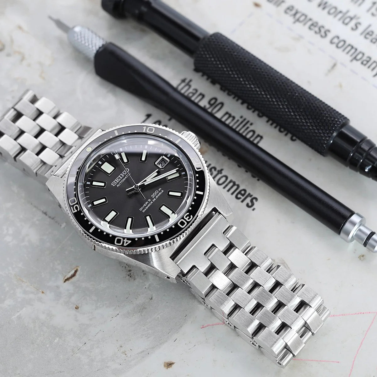 Strapcode Engineer bracelet with V-clasp attached to silver dive watch with black dial and bezel on desk with pens