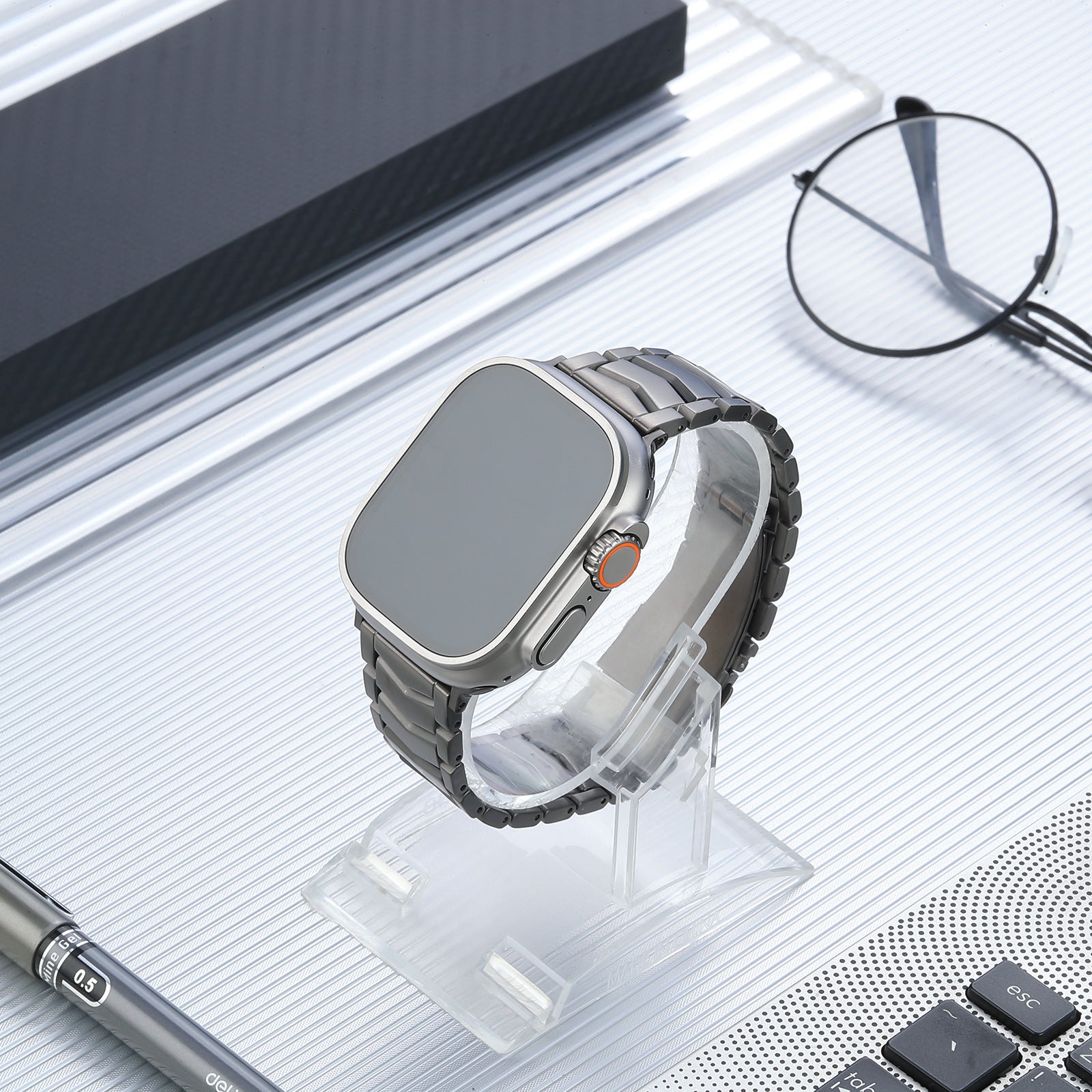 Titanium smartwatch with arched bracelet band and orange digital crown on clear display stand with glasses and tech accessories