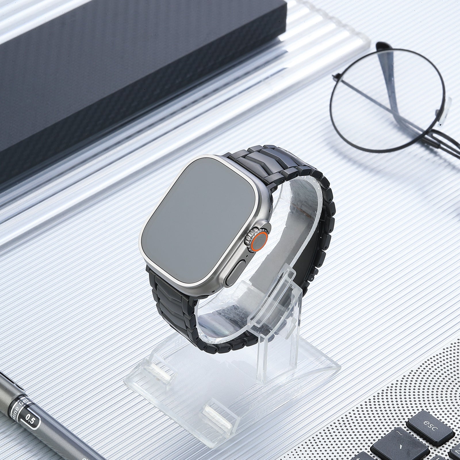 Titanium smartwatch with arched black bracelet and orange digital crown on clear display stand, styled flatlay with eyeglasses and tech accessories