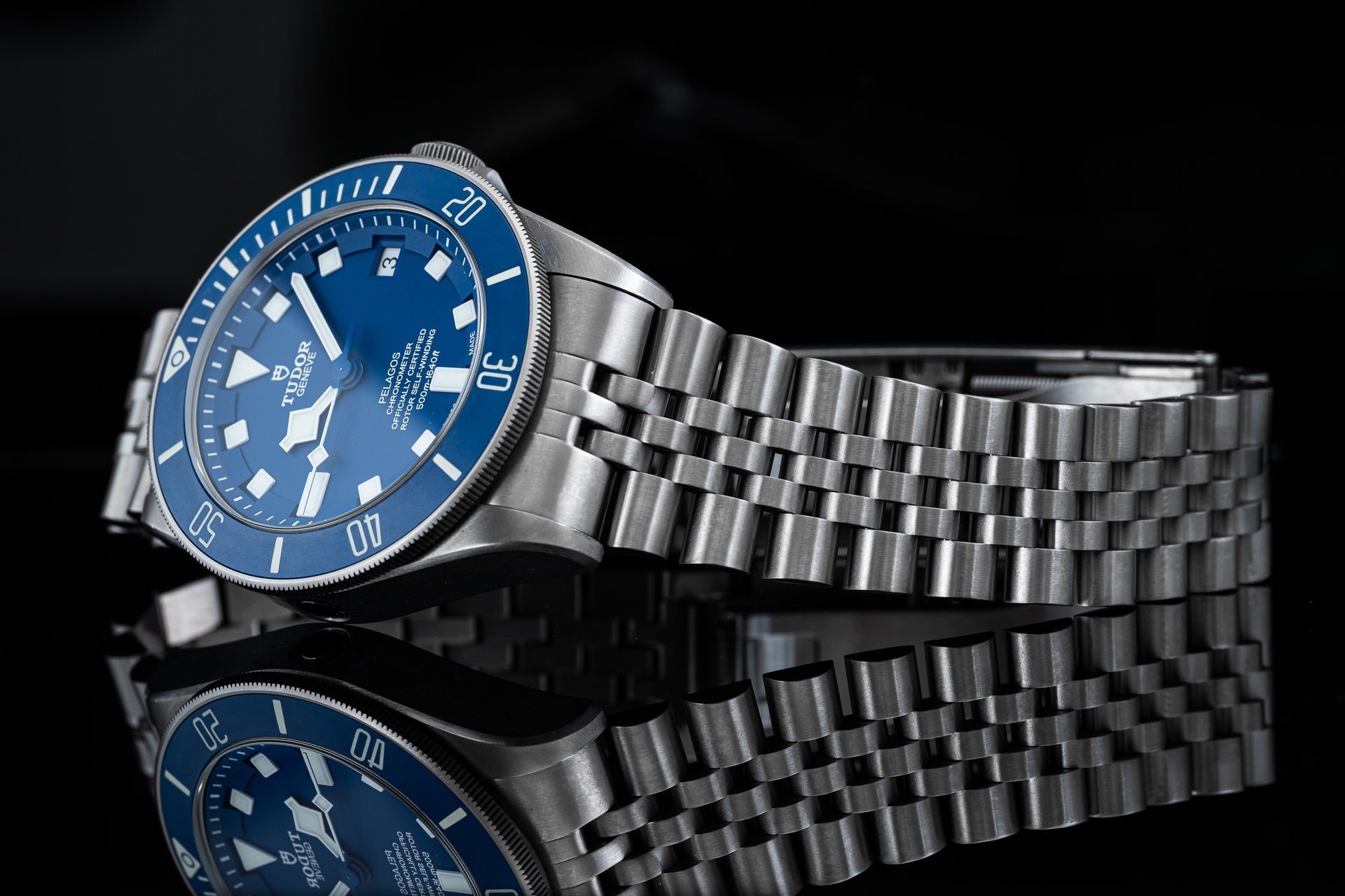 Stainless steel dive watch with blue dial and matching metal bracelet on black background with reflection