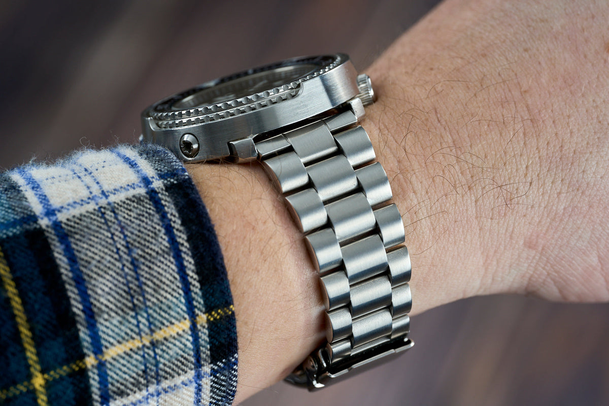 Stainless steel watch bracelet with straight end links on wrist wearing tartan fabric, showcasing polished metal construction and professional fit