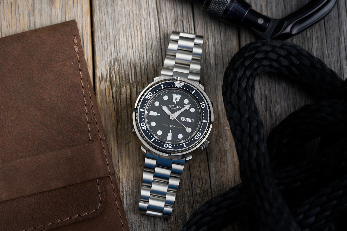 Seiko automatic dive watch with black dial and steel bracelet on wooden surface next to leather wallet and headphones