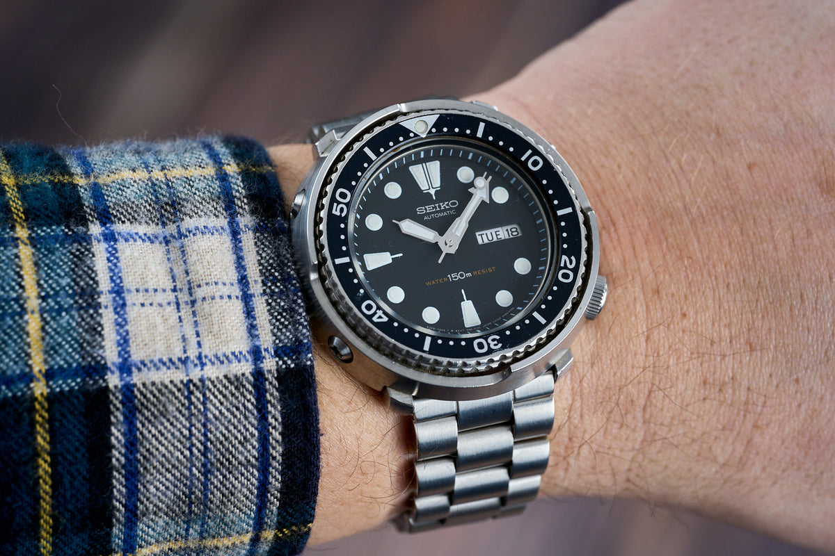 Seiko automatic dive watch with black dial, white hour markers, day-date window, stainless steel bracelet worn on wrist with blue plaid fabric cuff