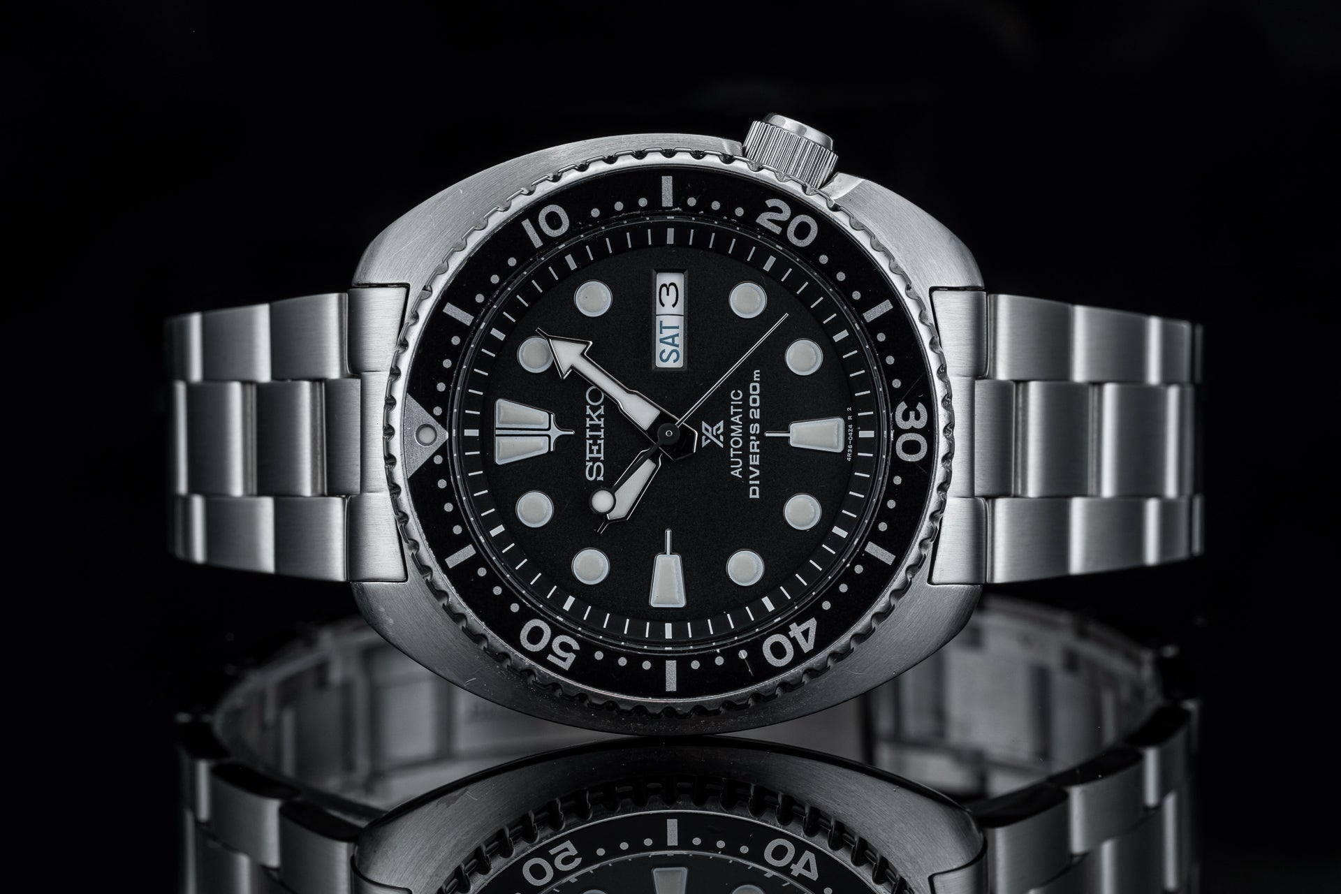 Seiko automatic diver watch with stainless steel bracelet, black dial, day-date window, and rotatable bezel on reflective surface
