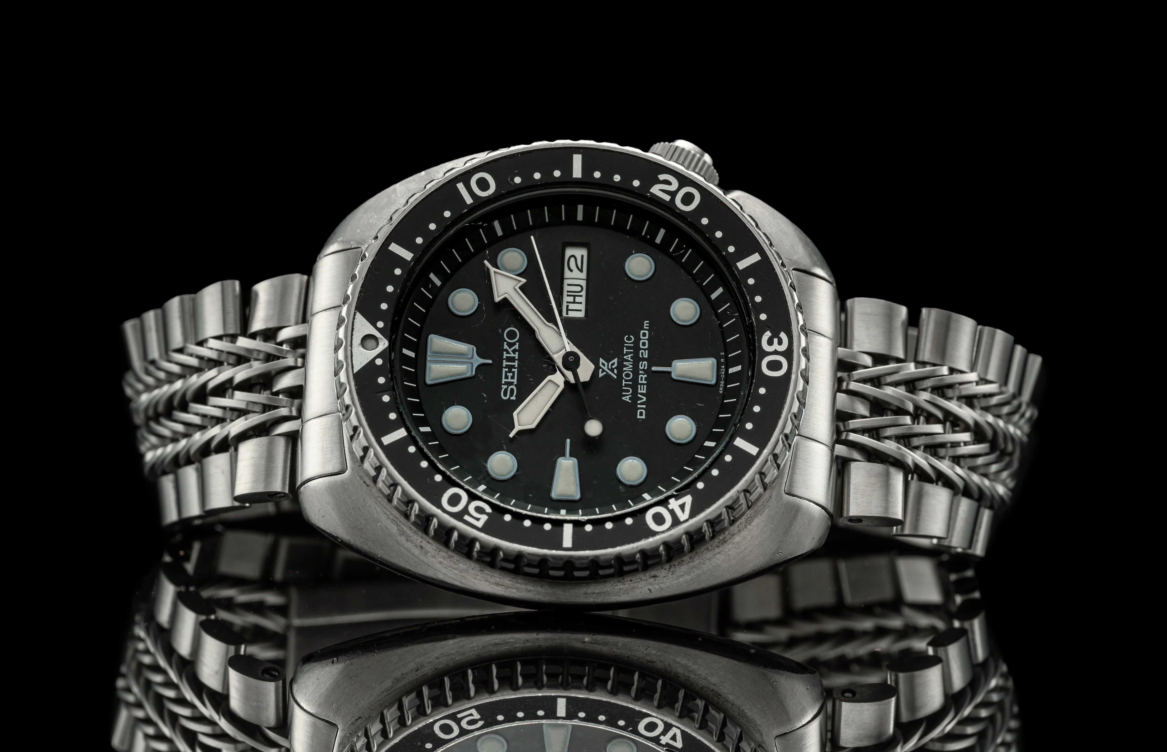 Seiko automatic dive watch with black dial, stainless steel bracelet, day-date window, luminous hour markers and hands on reflective surface