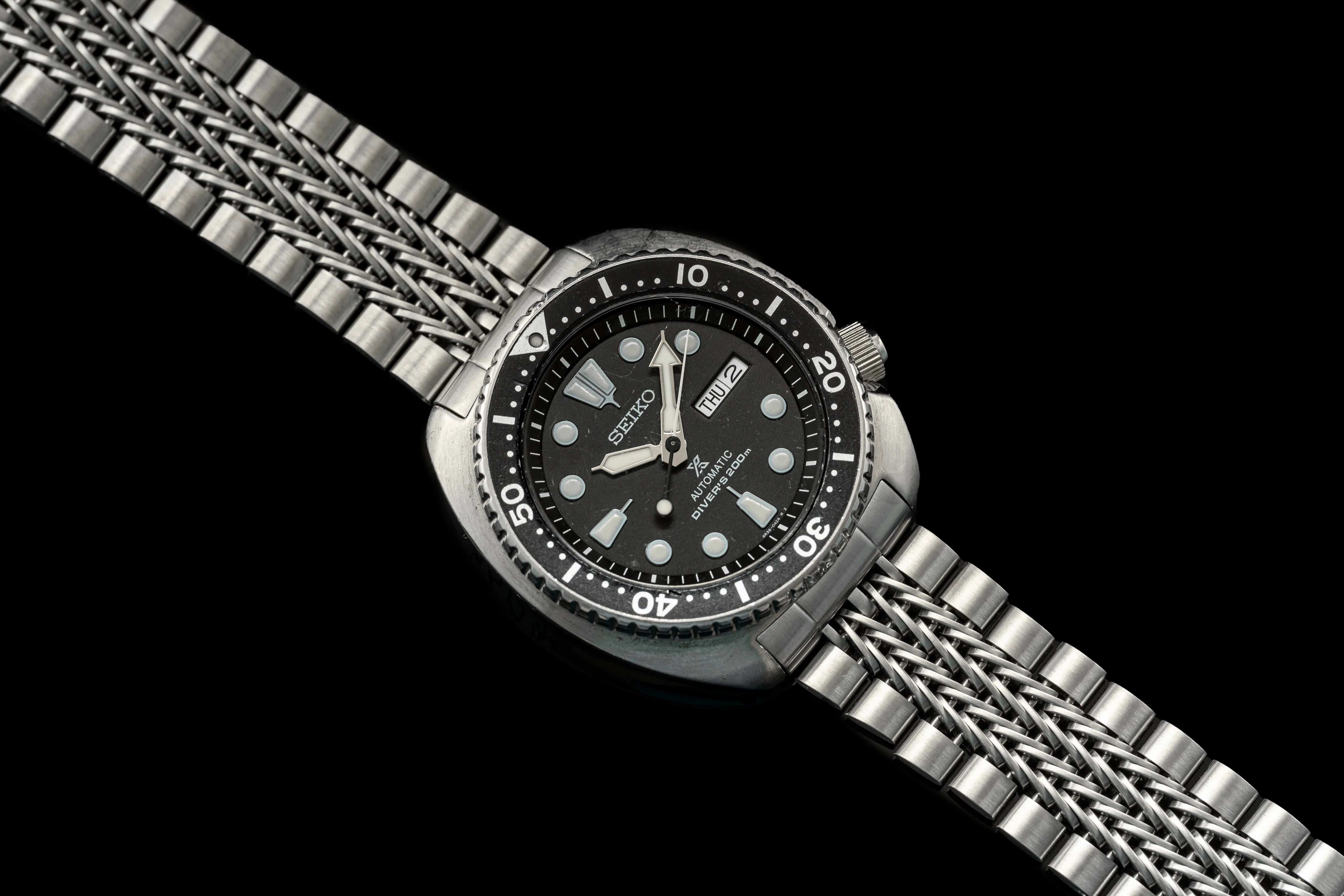 Seiko diver watch with black dial, stainless steel case, and textured mesh bracelet on black background