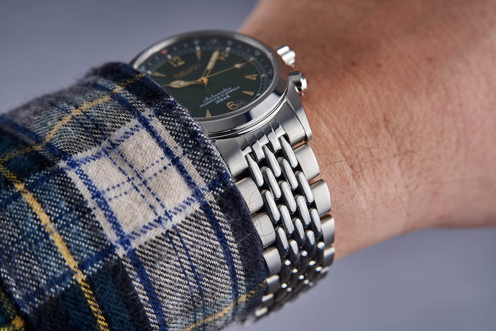 Stainless steel watch with black dial and gold markers on beaded bracelet worn over plaid shirt sleeve