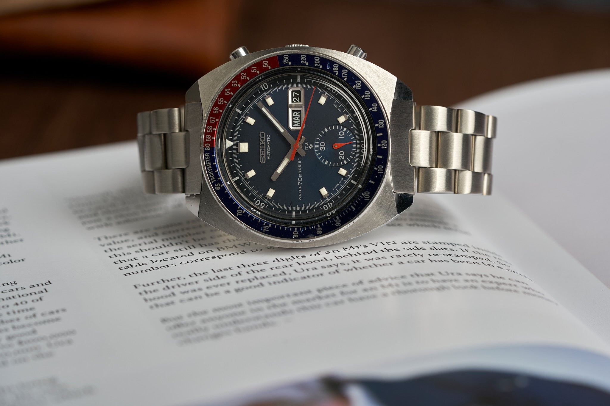 Vintage Seiko 6139 chronograph watch with stainless steel bracelet, black dial, day-date window, and red accents on book page