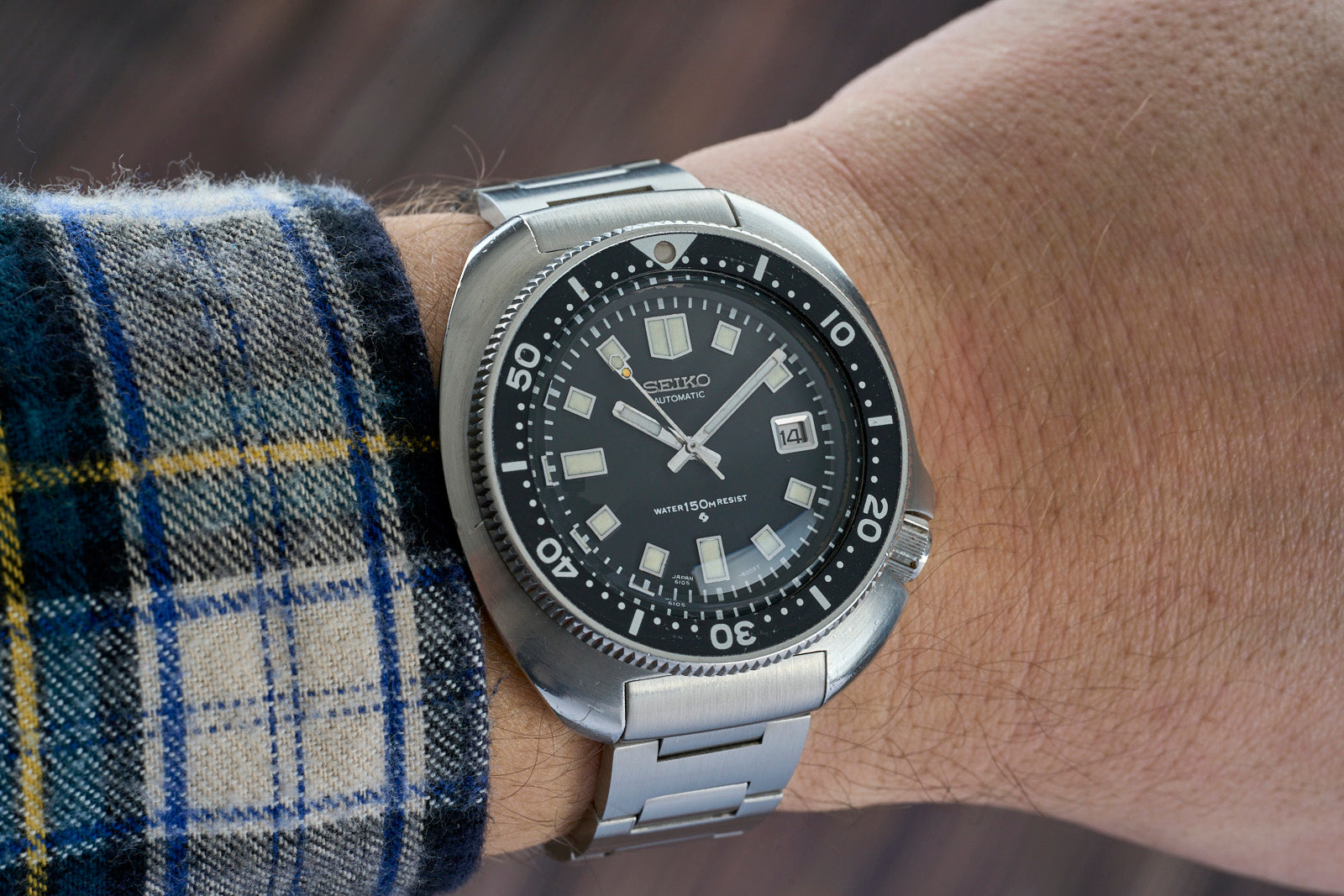 Seiko automatic dive watch with black dial, date window, and stainless steel bracelet worn on wrist with plaid shirt sleeve