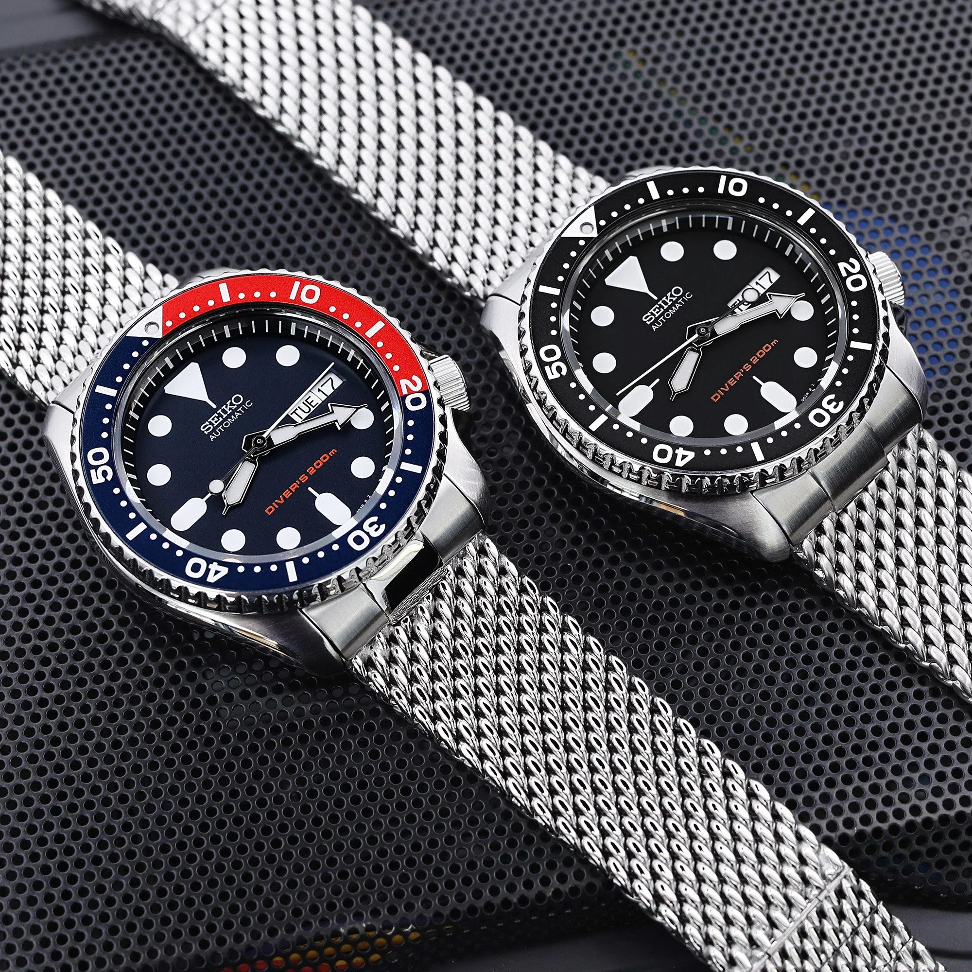 Two Seiko dive watches with stainless steel mesh bracelets on textured background, one with red and blue bezel, one with black bezel