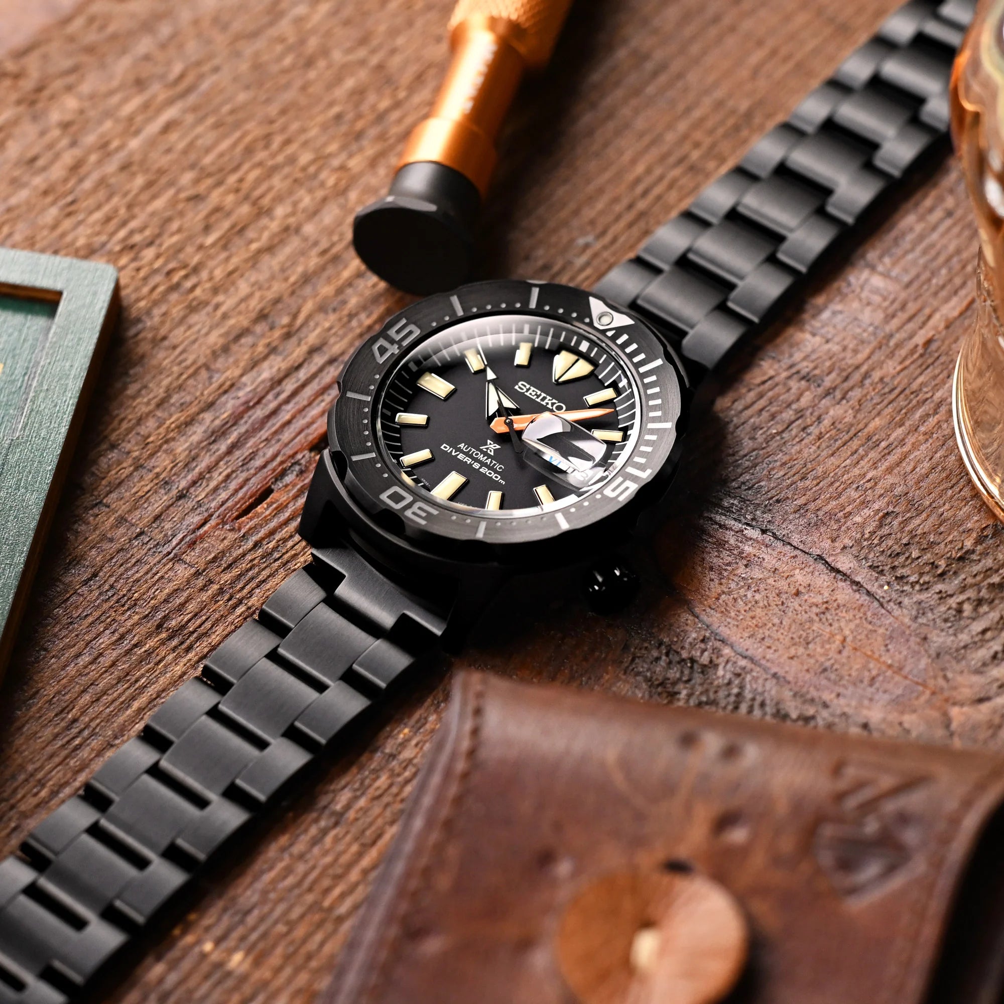 Black stainless steel watch bracelet with V-clasp and DLC coating displayed on wooden surface with accessories