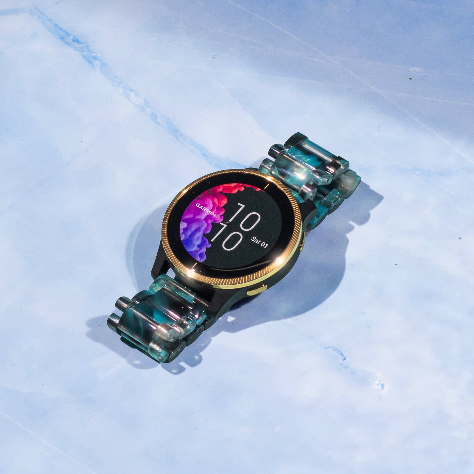 Garmin smartwatch with gold bezel and teal crystal acetate band displaying time 10:10 on black AMOLED screen with pink purple ink effect wallpaper