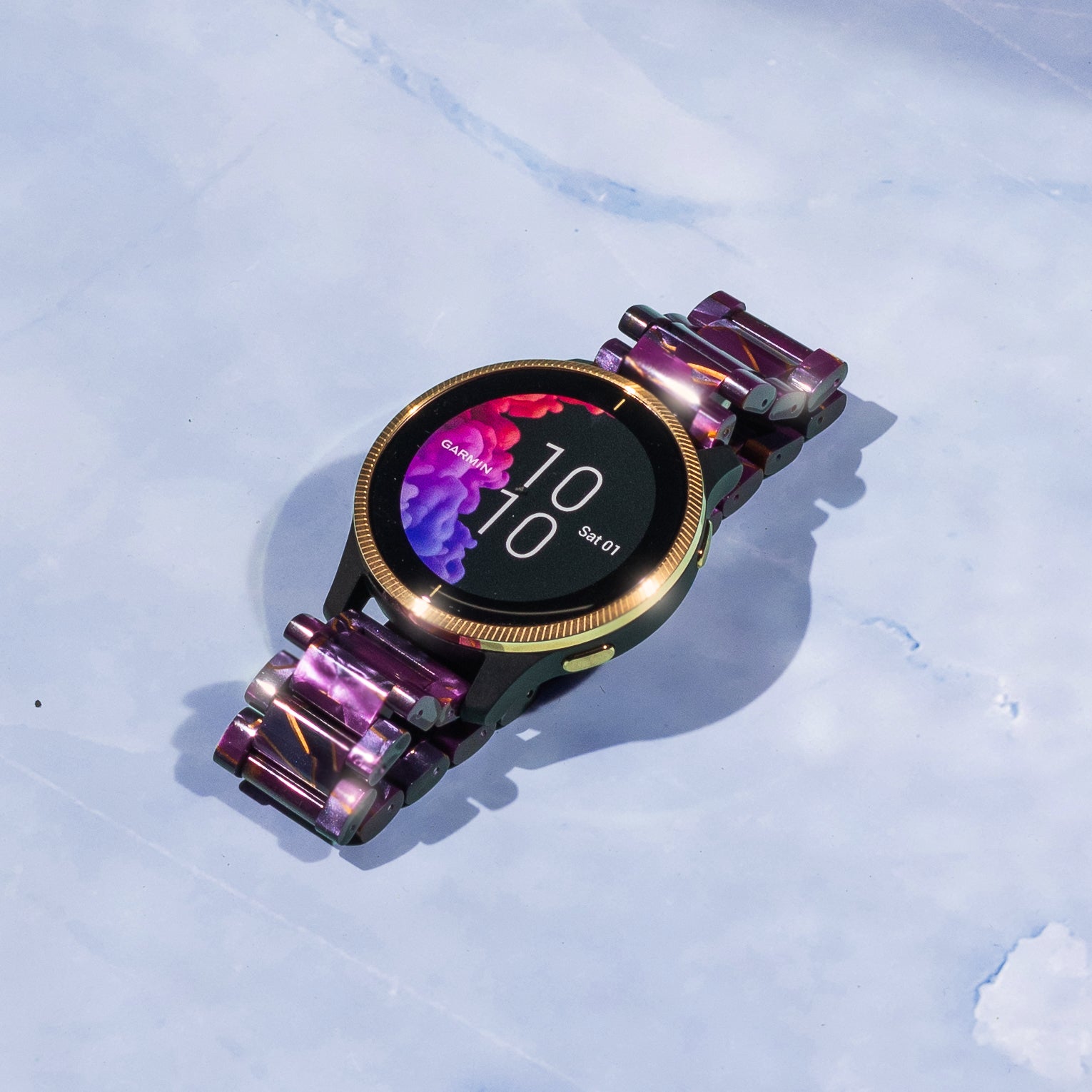 Garmin smartwatch with purple crystal acetate quick-release band, gold bezel, and digital display showing 10:10 time on white background