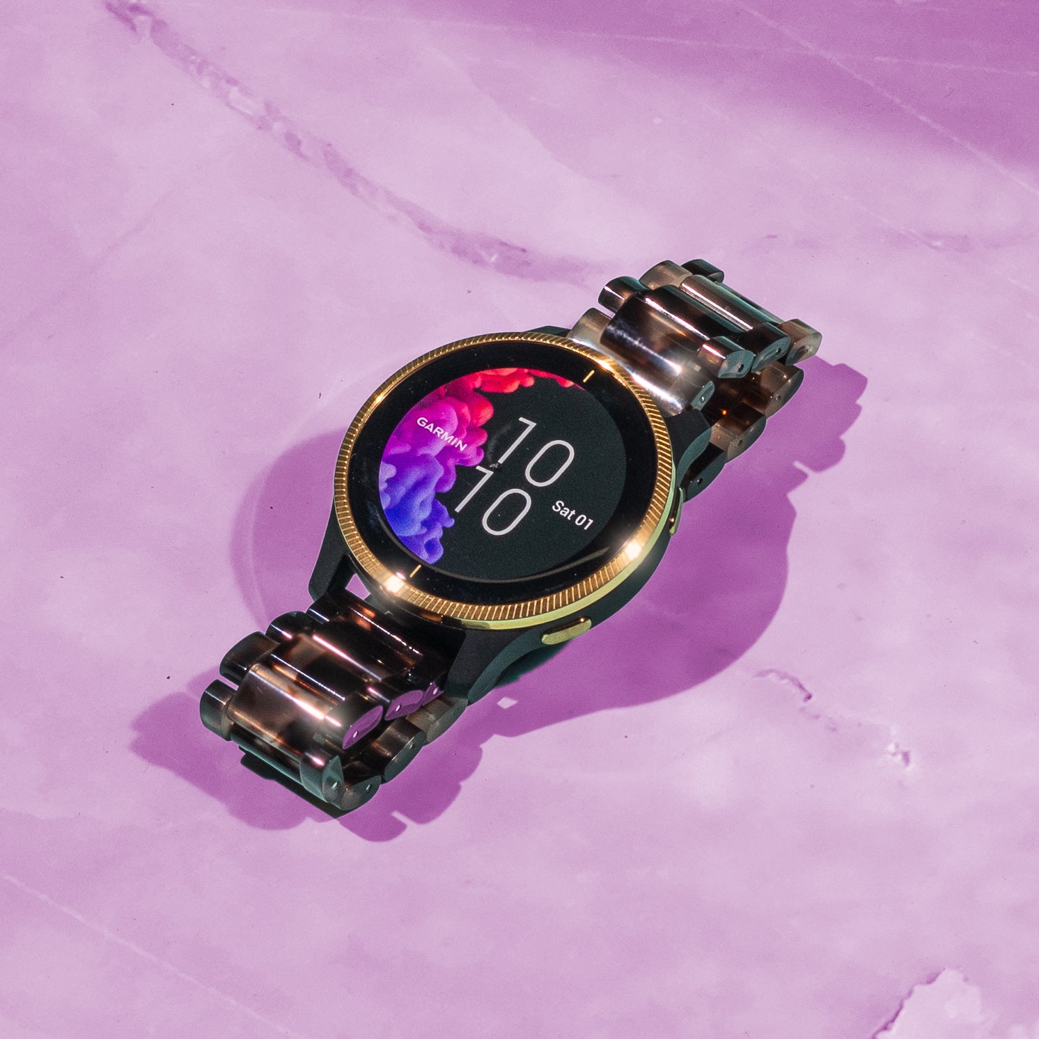 Garmin smartwatch with tortoiseshell dark brown acetate band and gold bezel on pink background displaying time