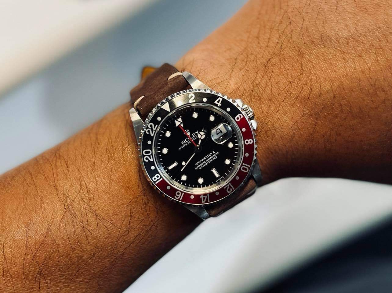 Luxury sport watch with black dial, red and black rotating bezel, date window, and brown leather strap worn on wrist