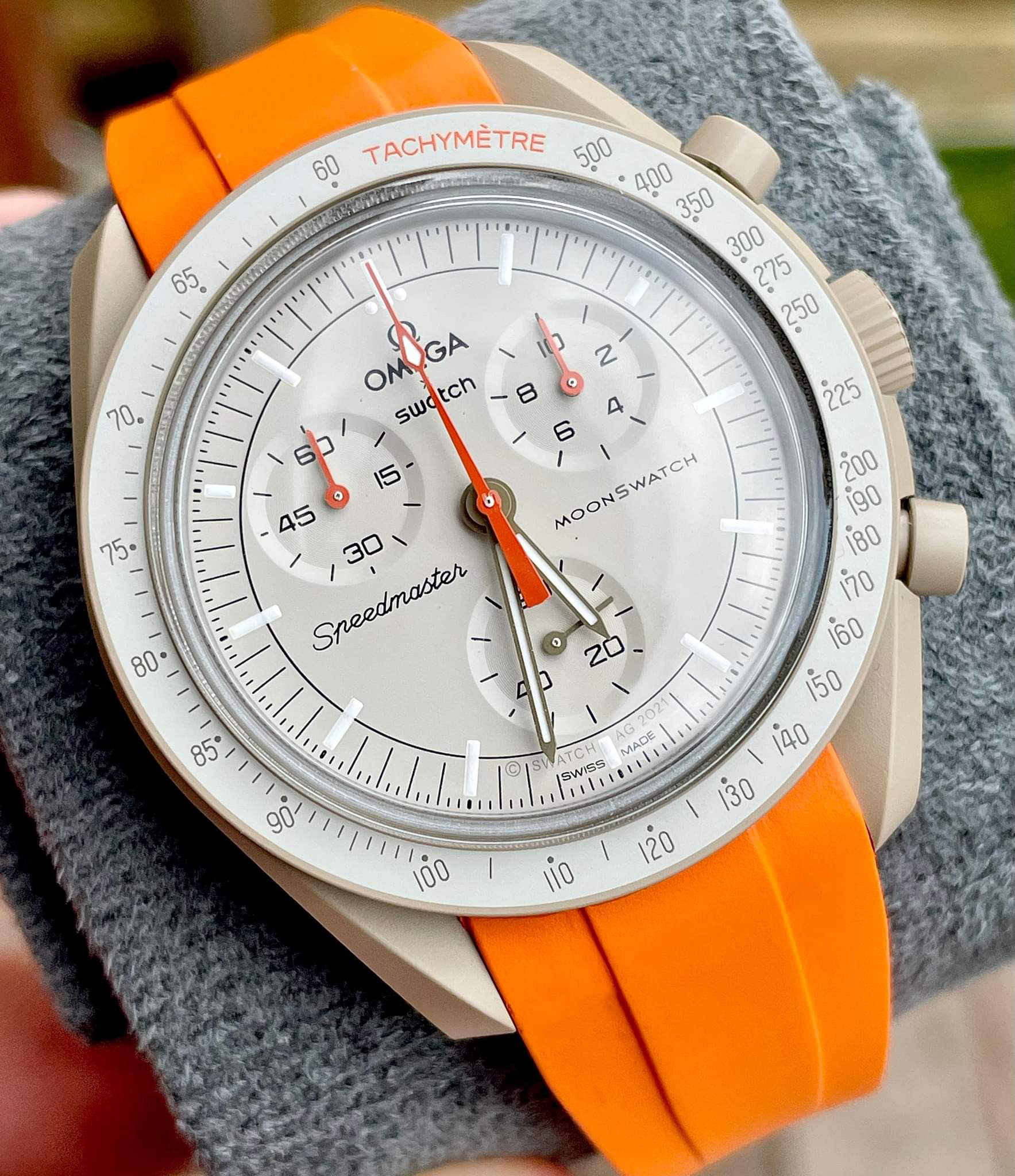 Omega Swatch MoonSwatch Speedmaster chronograph watch with white dial, orange rubber strap and tachymetre bezel on gray fabric background