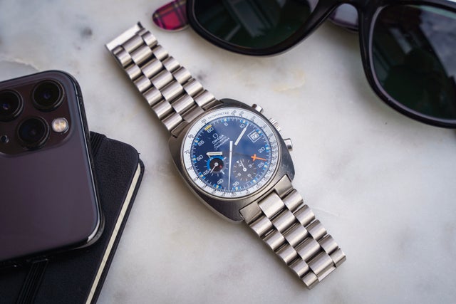 Silver stainless steel chronograph watch with blue dial and metal bracelet, displayed with smartphone and sunglasses