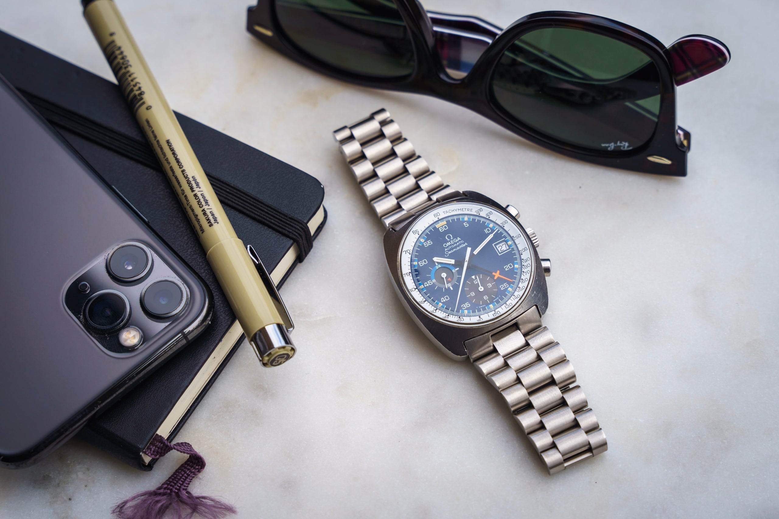 Omega Seamaster chronograph watch with stainless steel bracelet and blue dial, displayed with smartphone, sunglasses, pen and notebook