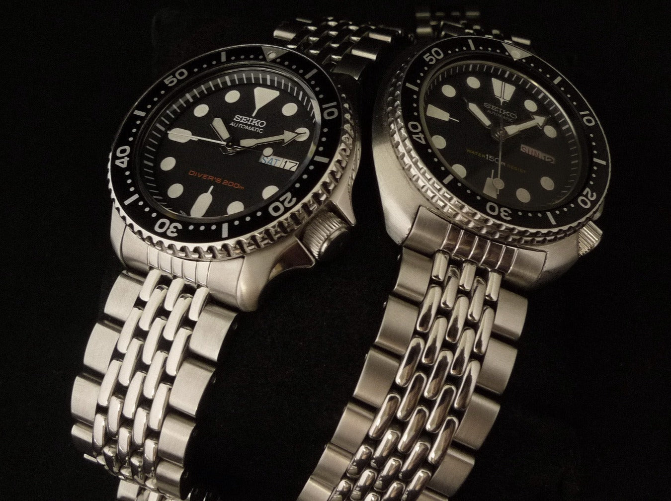 Two Seiko automatic dive watches with black dials, luminous hands, stainless steel beads of rice bracelets, and rotating bezels on black background