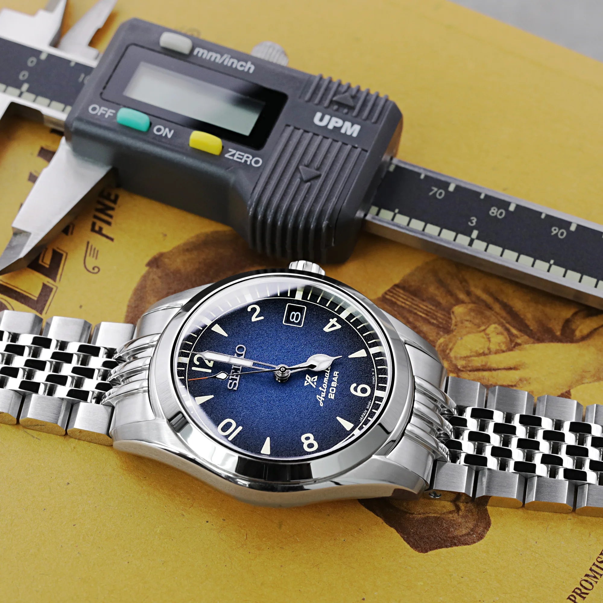 Strapcode Asteroid 20mm stainless steel watch bracelet on Seiko Alpinist with digital caliper and measuring scale on yellow background
