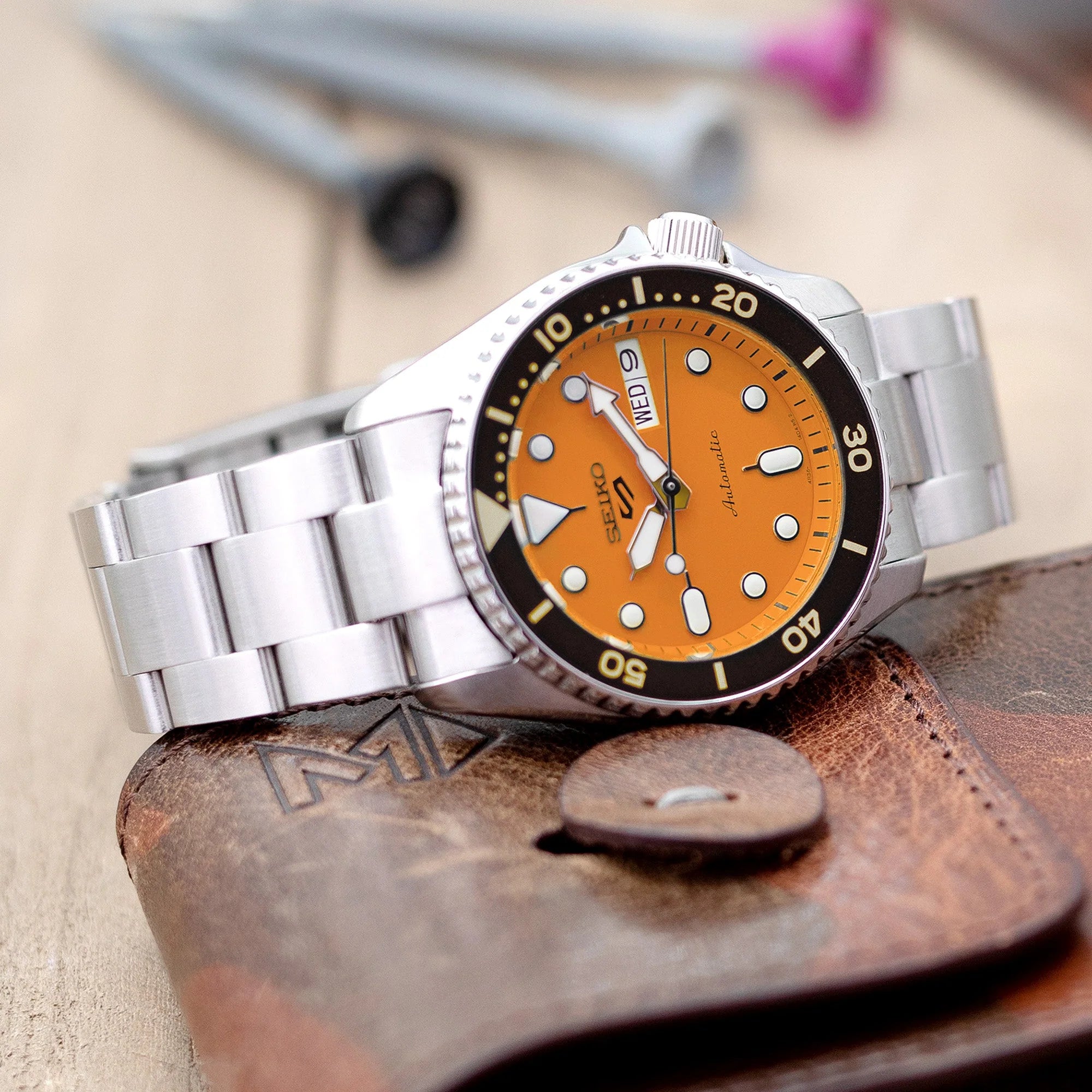 Silver stainless steel Seiko 5 Sports watch with orange dial and rotating bezel on matching metal bracelet