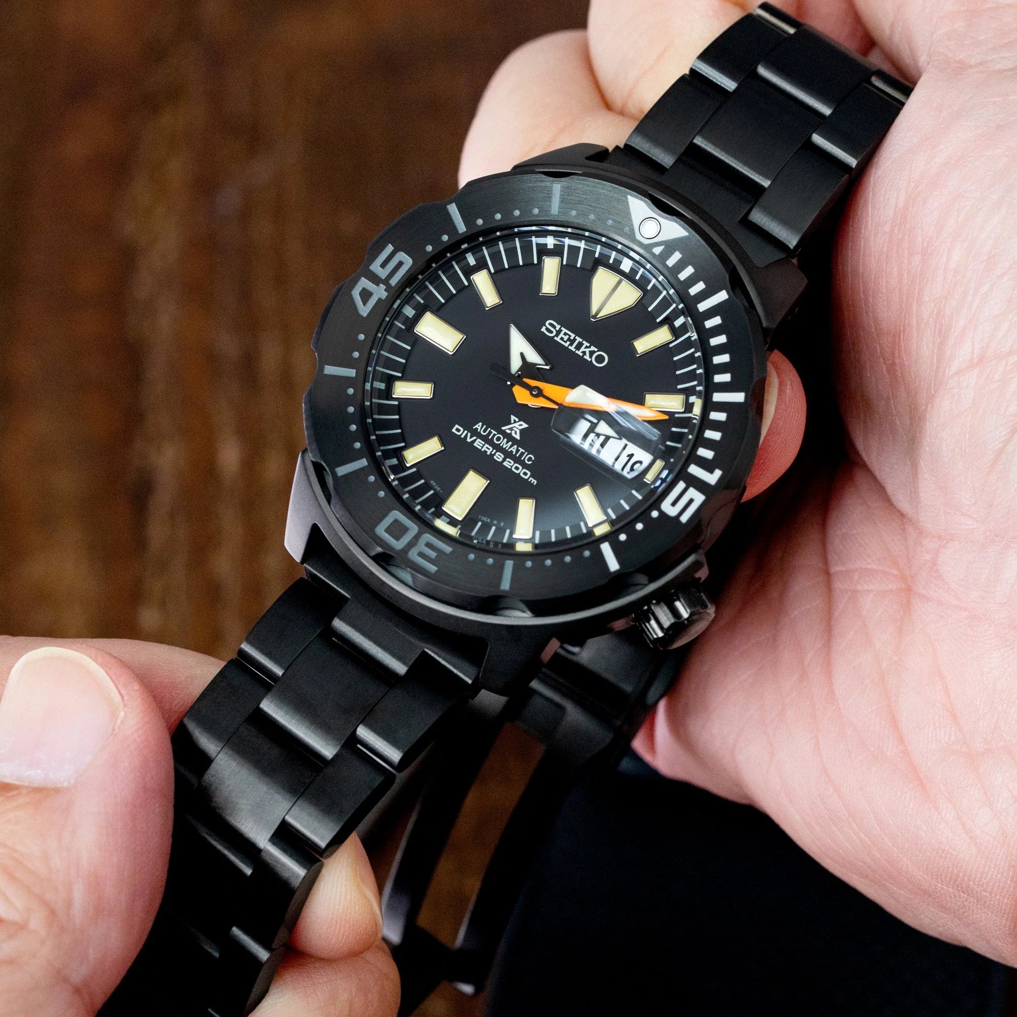 Black Seiko automatic diver watch with luminous markers and date window, held in hand displaying steel bracelet and DLC-coated case