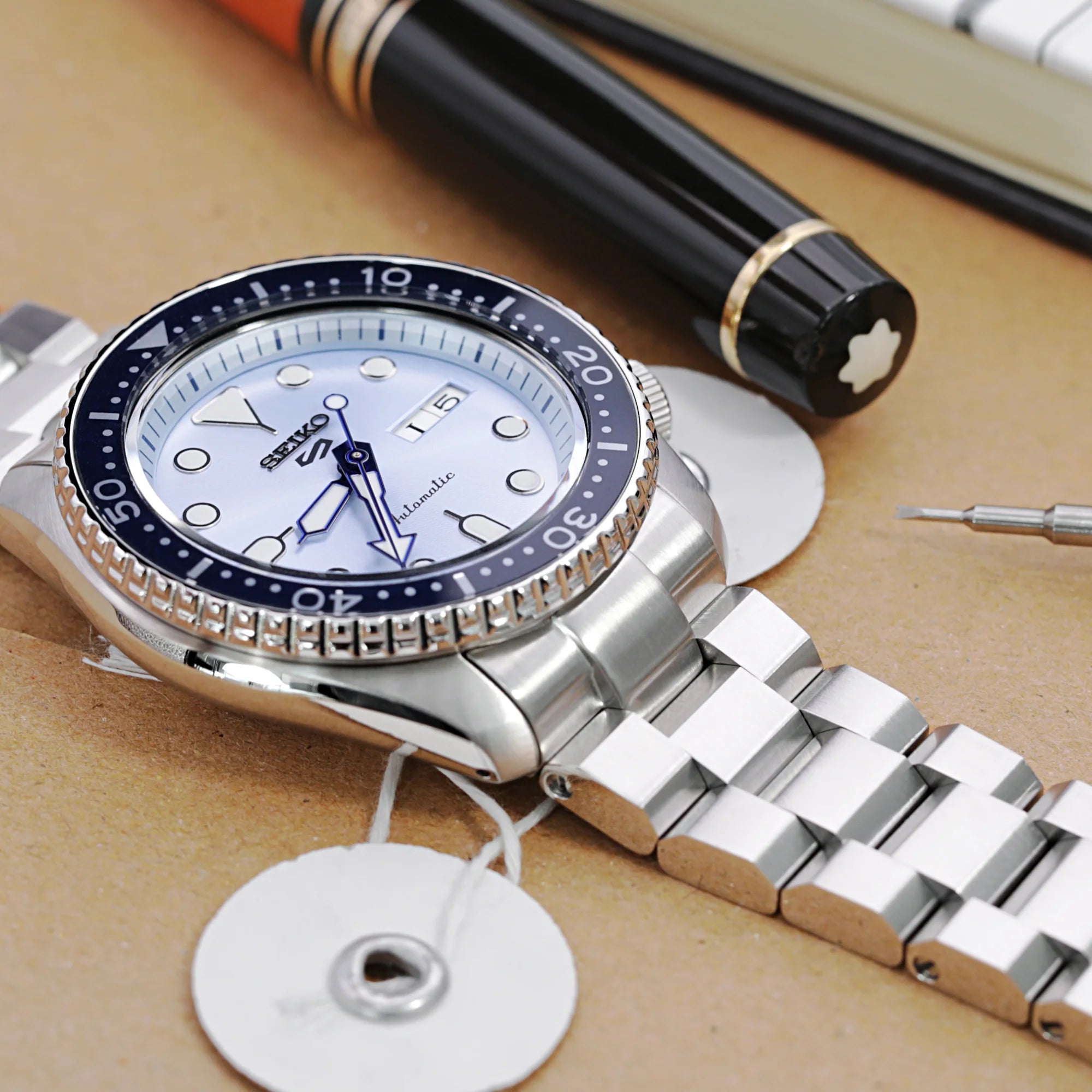 Seiko 5 automatic watch with stainless steel Hexad bracelet on tan background with pens