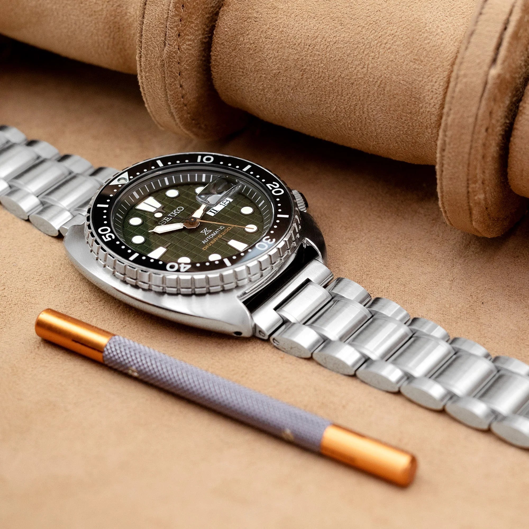 Stainless steel dive watch with green dial and metal bracelet featuring V-clasp, styled with brown leather strap and tool on tan background