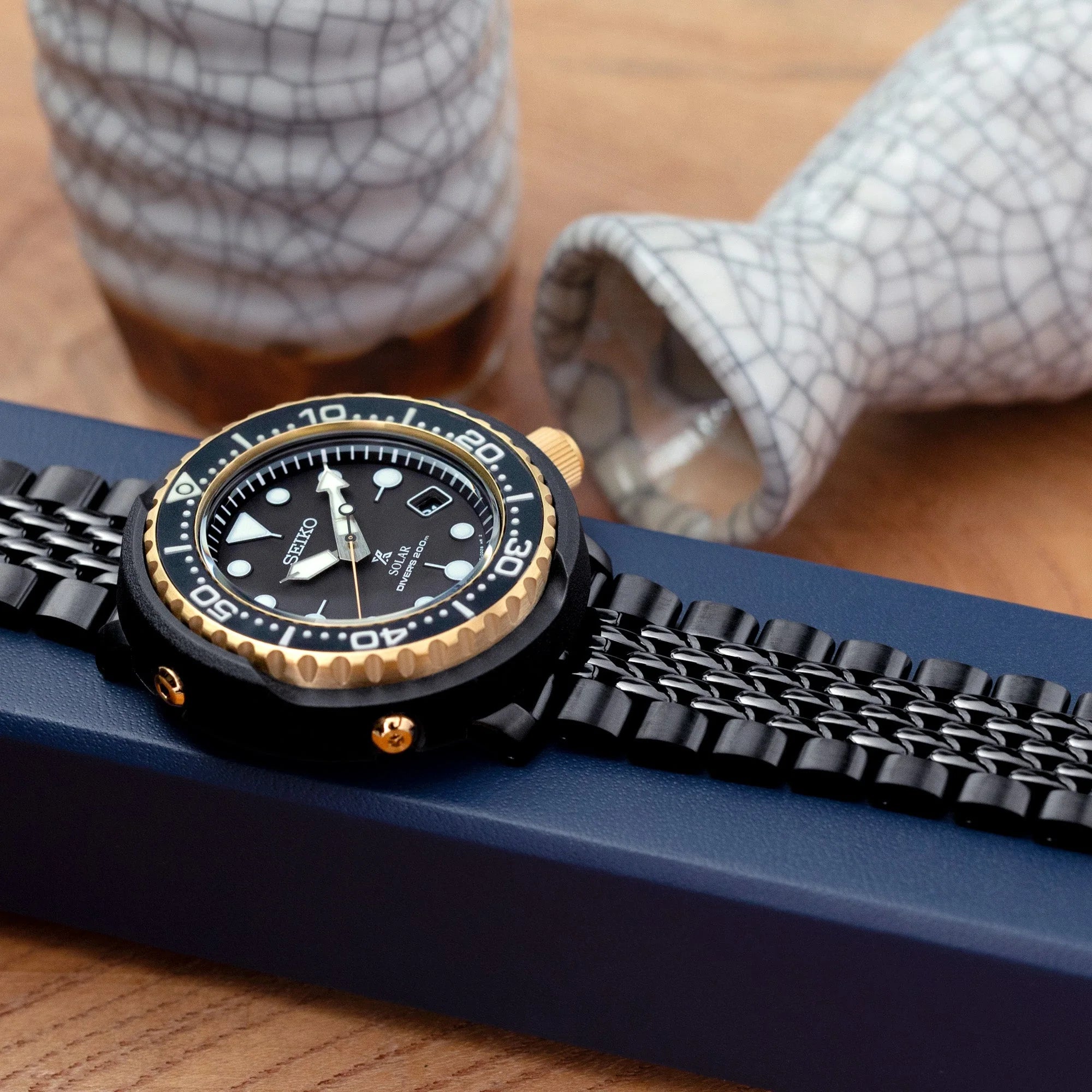 Black stainless steel watch bracelet with DLC coating, V-clasp, and gold accents on blue display box
