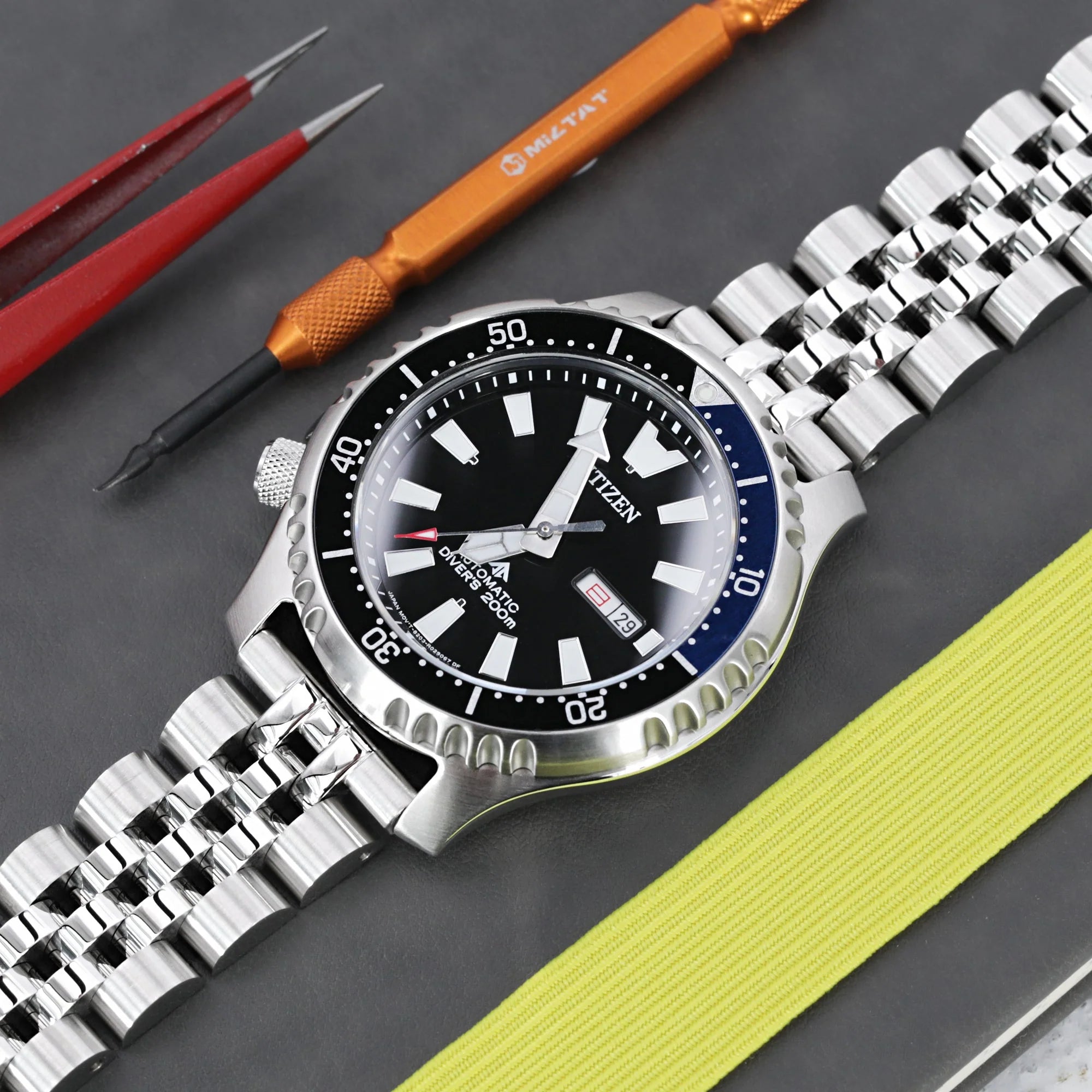Strapcode Angus-J Louis steel bracelet with V-clasp on dive watch with black dial, featuring precision watchmaking tools and yellow strap