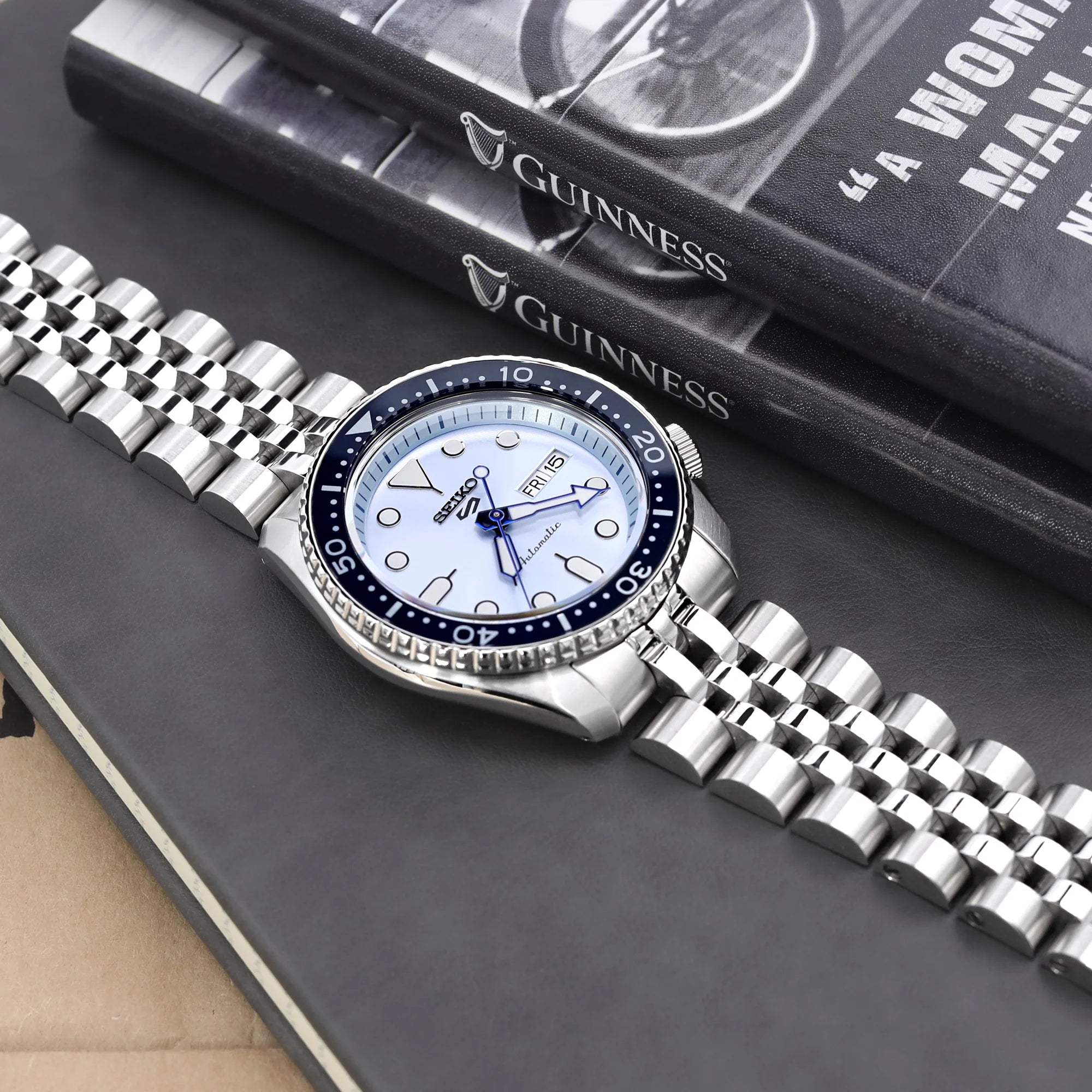 Seiko 5 watch with white dial and steel jubilee bracelet on black surface next to Guinness books