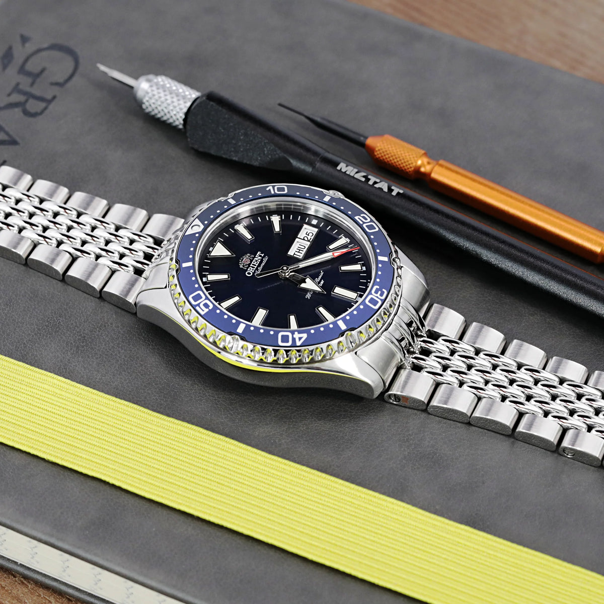 Silver stainless steel dive watch with blue and yellow bezel, black dial, and matching bracelet on gray surface with repair tools