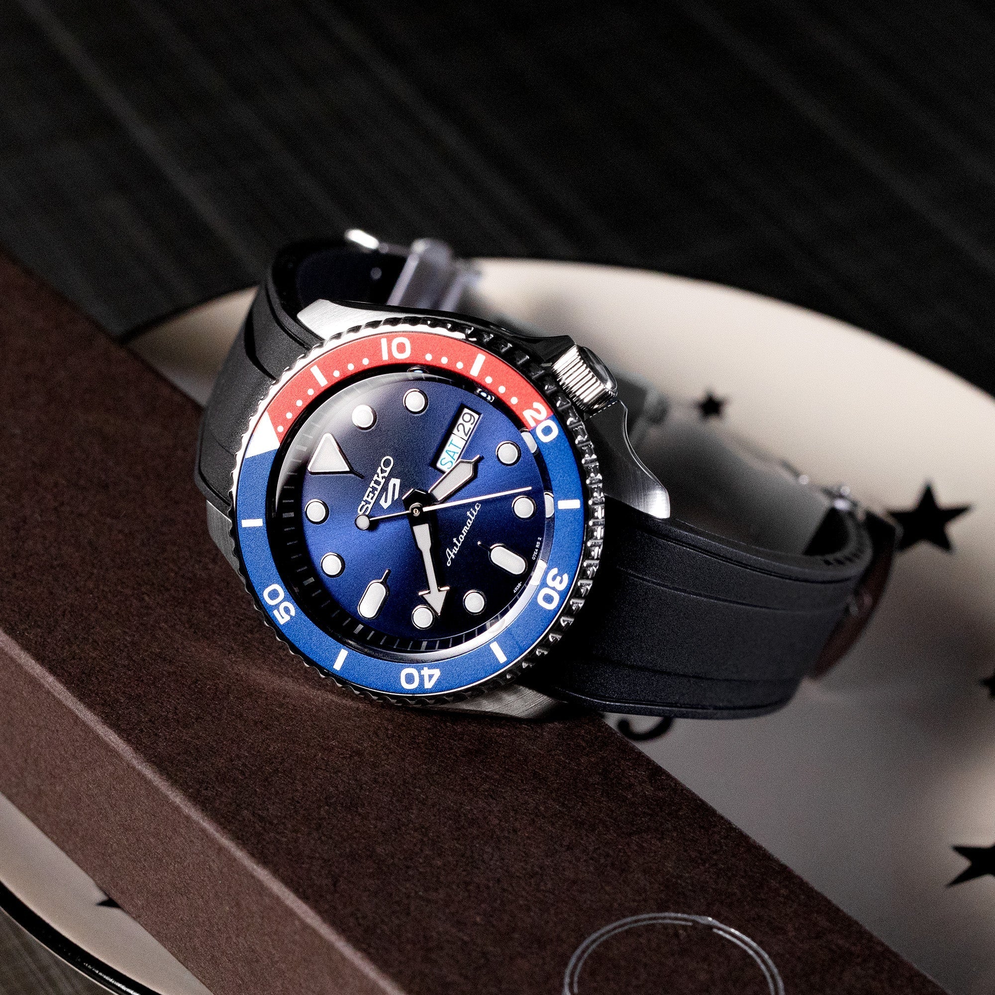 Seiko 5 Sports dive watch with blue and red bezel on black rubber strap with curved ends, displayed on brown and white surface