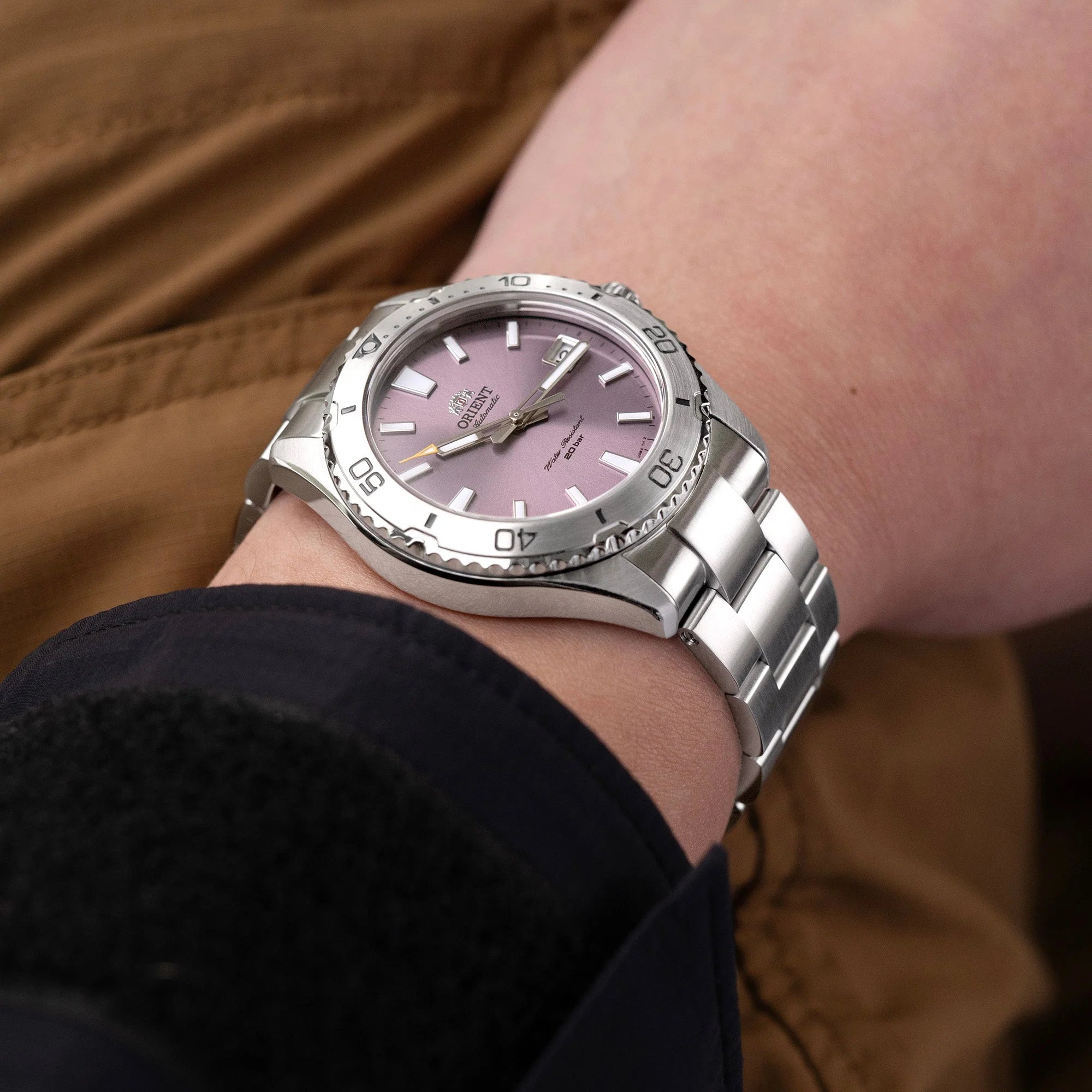 Silver stainless steel watch with mauve dial and rotating bezel worn on wrist with brown leather jacket