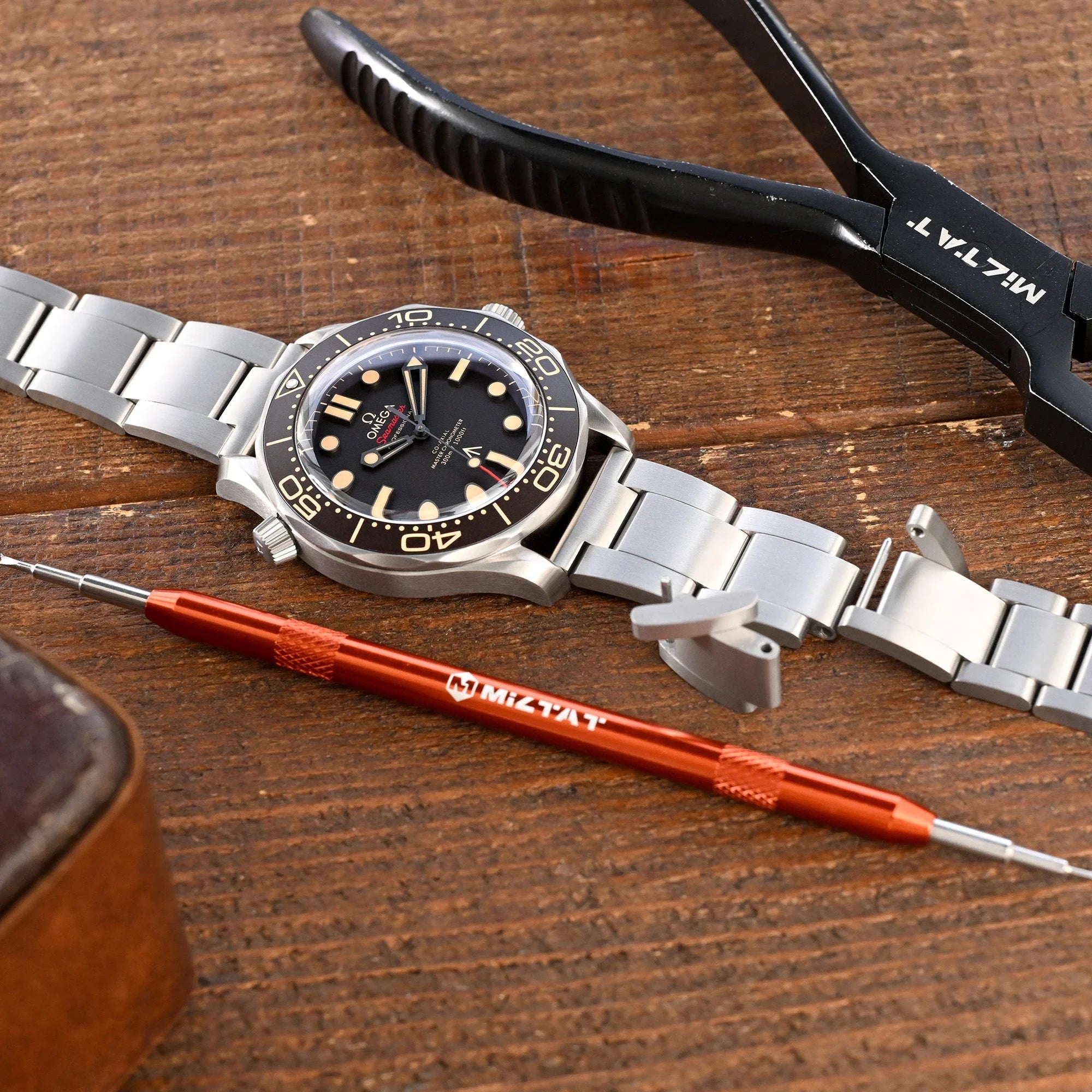 Stainless steel dive watch with black dial and gold hour markers next to titanium quick release watch band and watchmaking tools on wooden surface