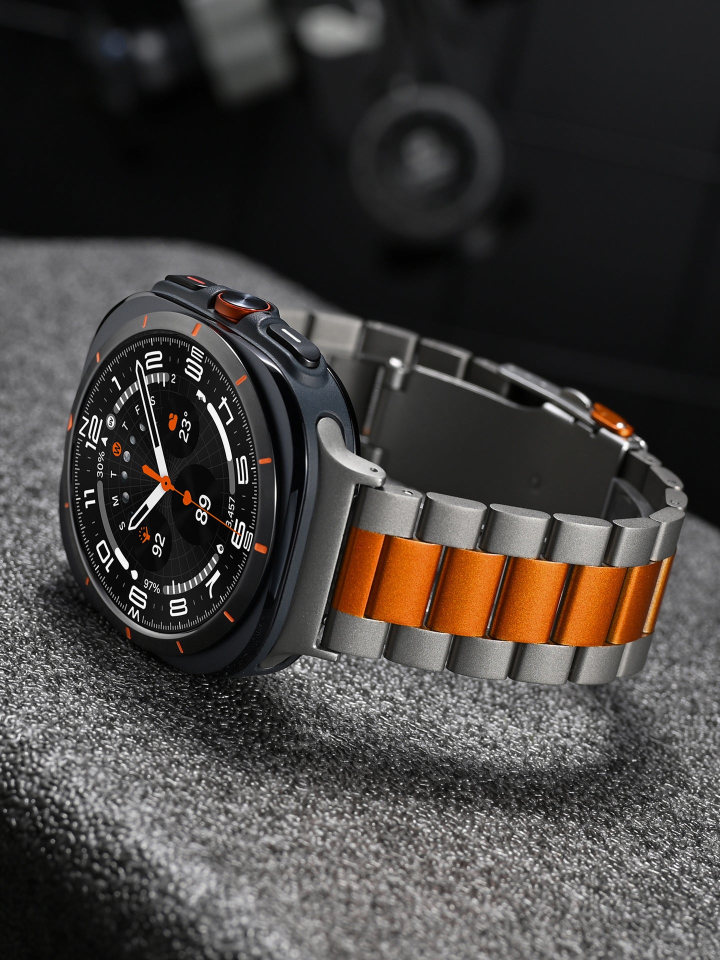 Black smartwatch with orange and silver steel bracelet, analog dial, chronograph subdials, and orange accents on gray surface