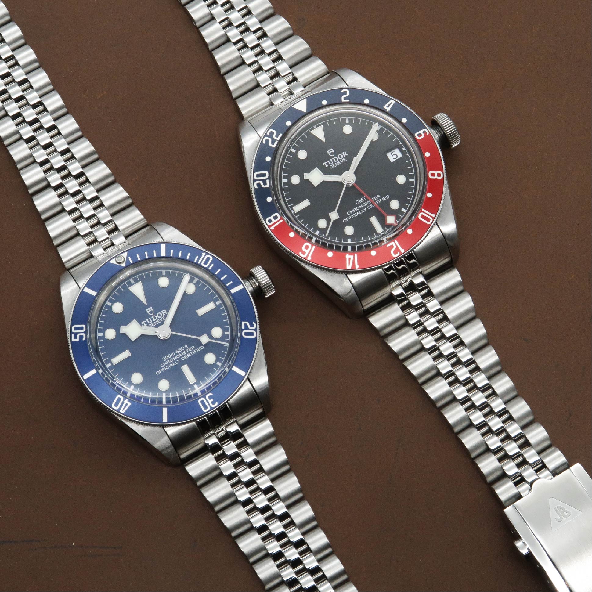 Two Tudor Black Bay men's dive watches with steel bracelets, blue and red bezels on brown background