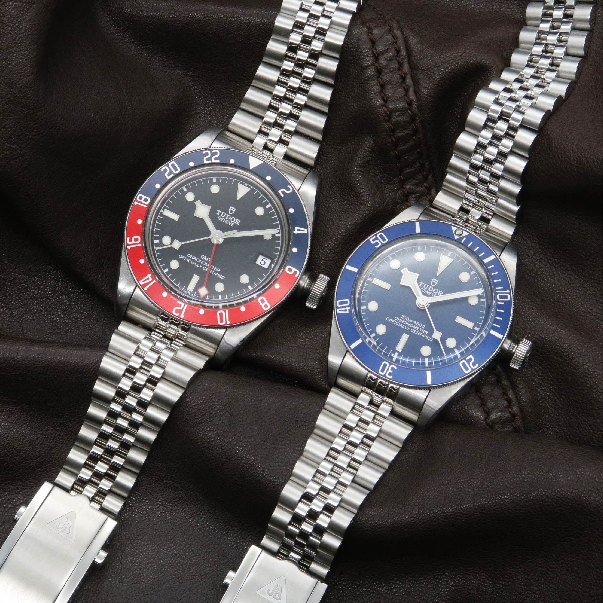 Two Tudor Black Bay steel watches with red-blue and blue bezels on jubilee bracelets displayed on black leather
