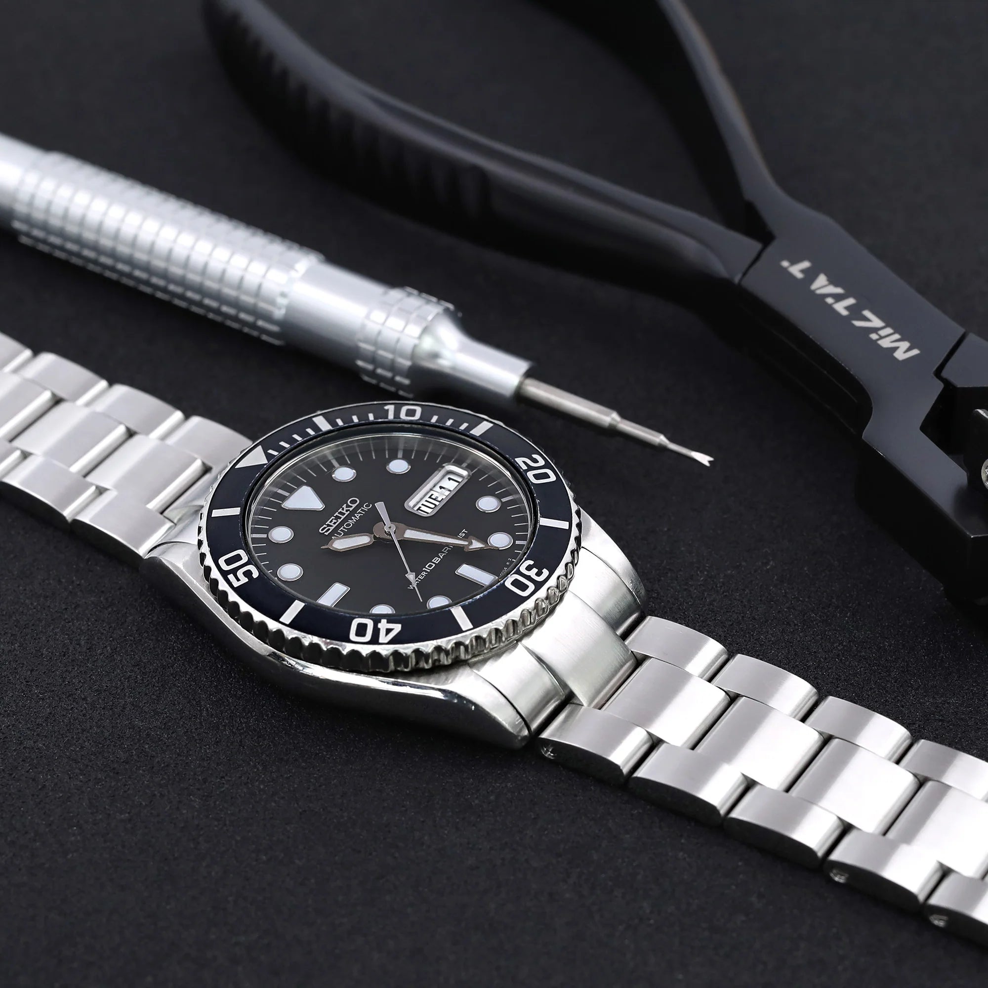 Seiko SKX023 dive watch with black dial and stainless steel Super-O Boyer bracelet on dark background with pen and tools