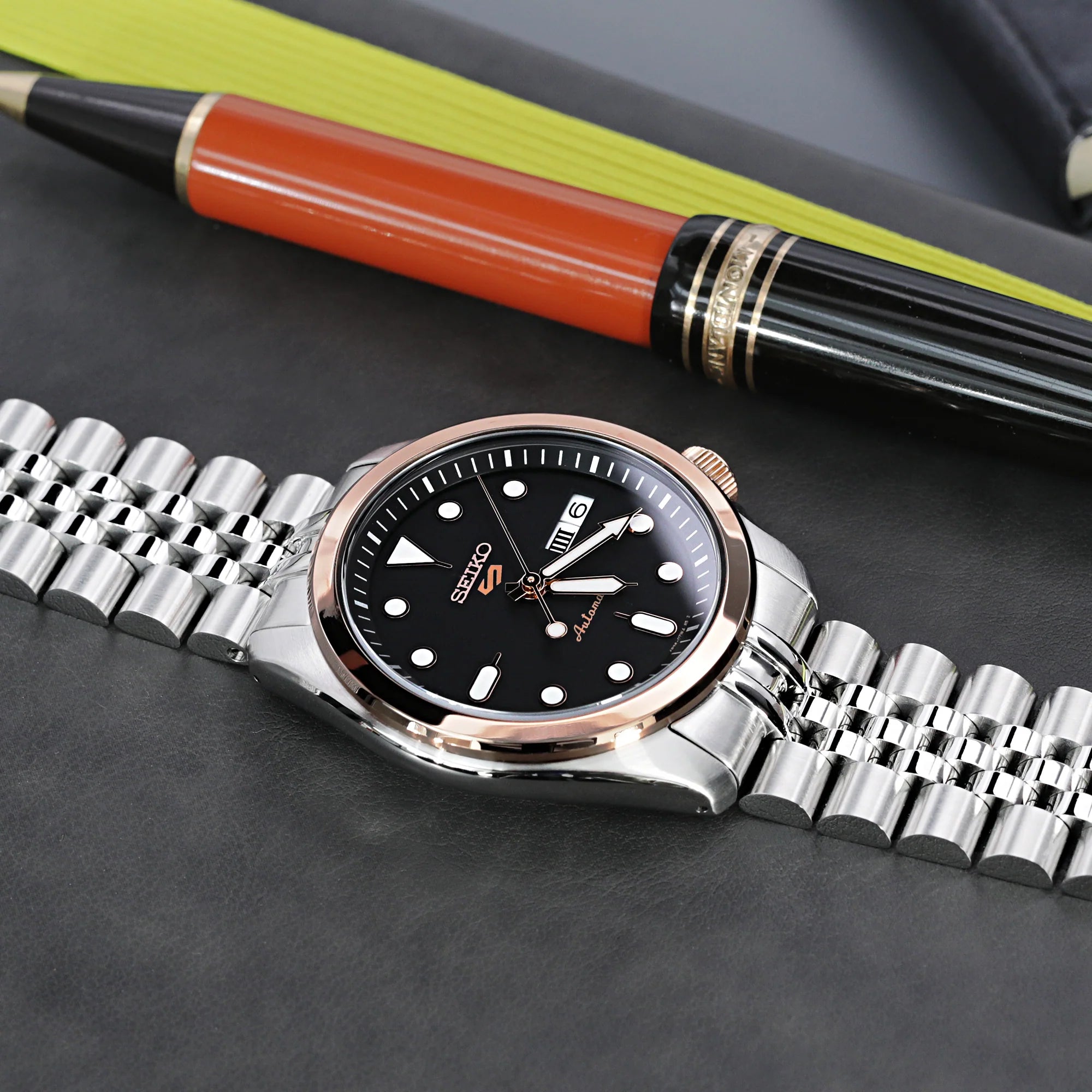 Seiko 5 automatic watch with stainless steel jubilee bracelet, black dial, and rose gold bezel next to luxury pen