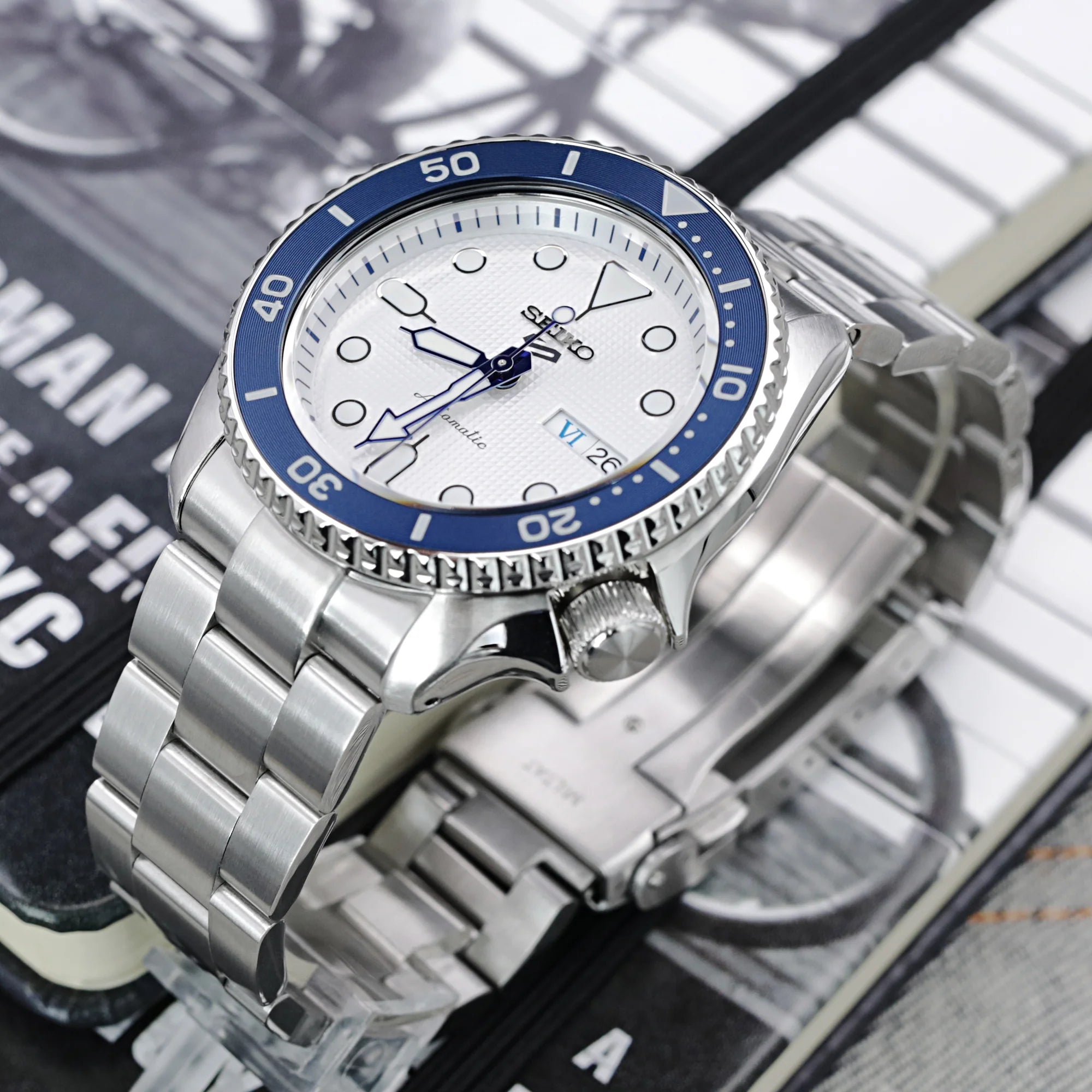 Stainless steel Seiko 5 dive watch with blue bezel, white dial, and Super-O Boyer bracelet on magazine background