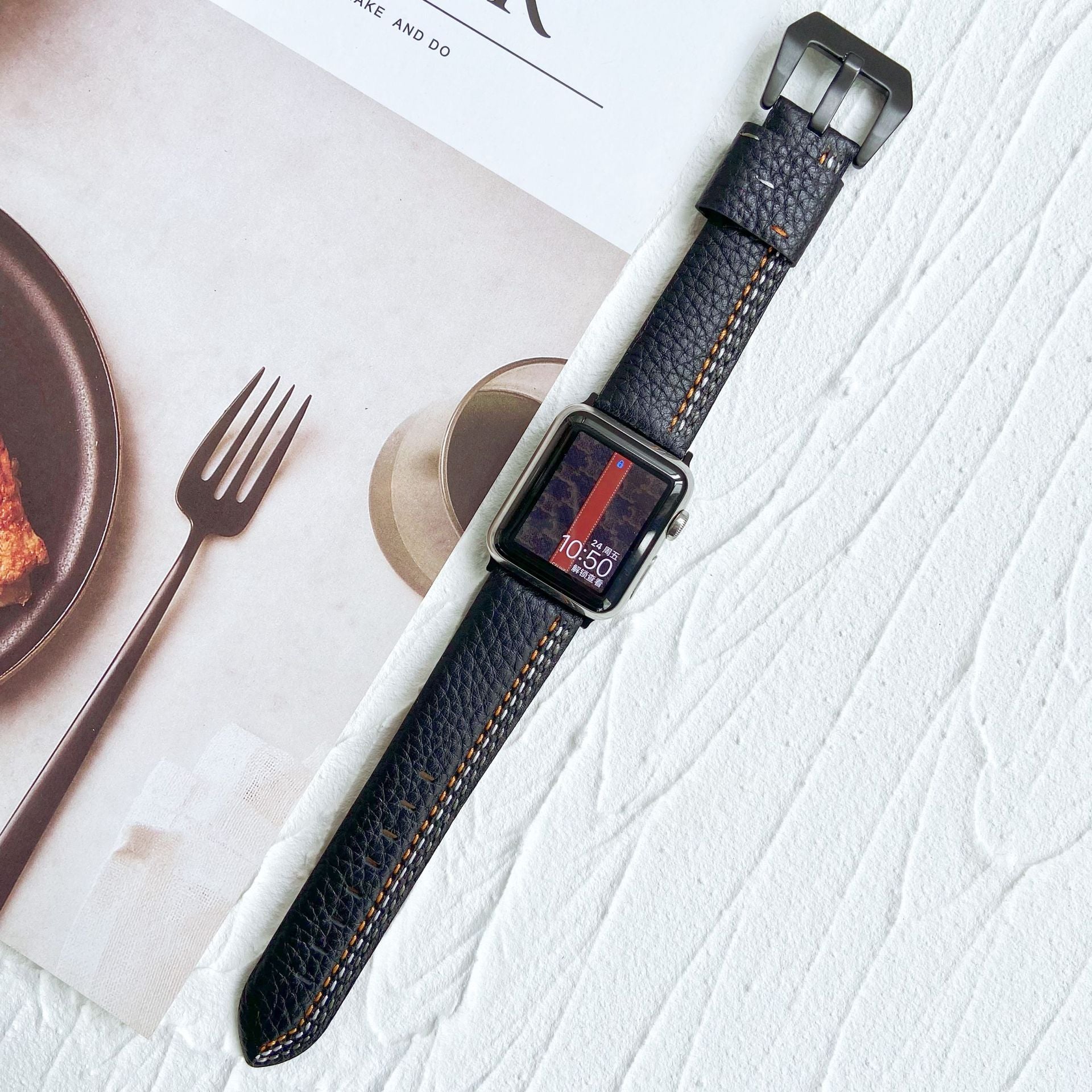 Black leather Apple Watch band with asymmetrical orange stitching, stainless steel buckle, displayed on white textured surface with dining setup
