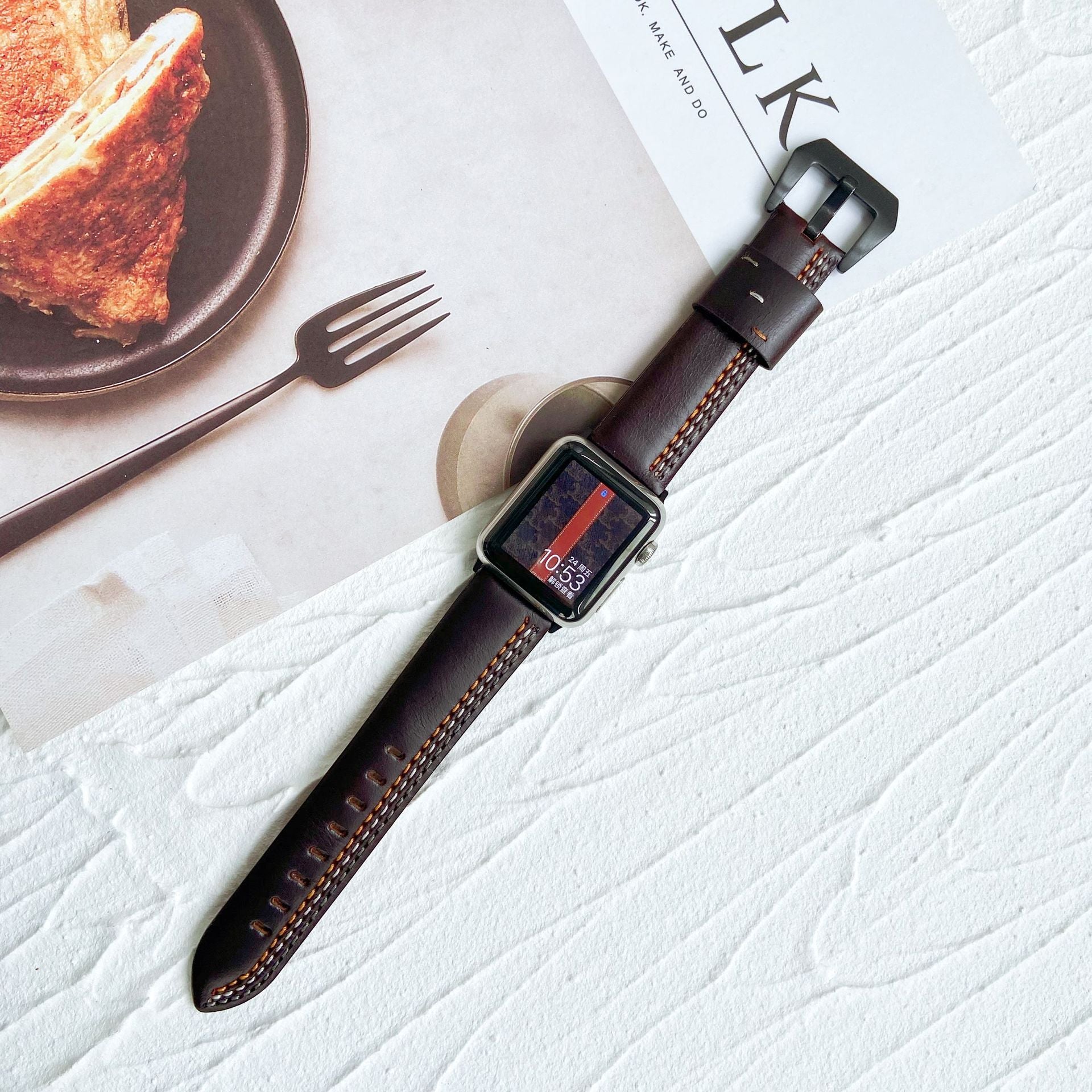 Dark brown leather Apple Watch band with asymmetrical stitching, displayed on white textured surface with dining scene