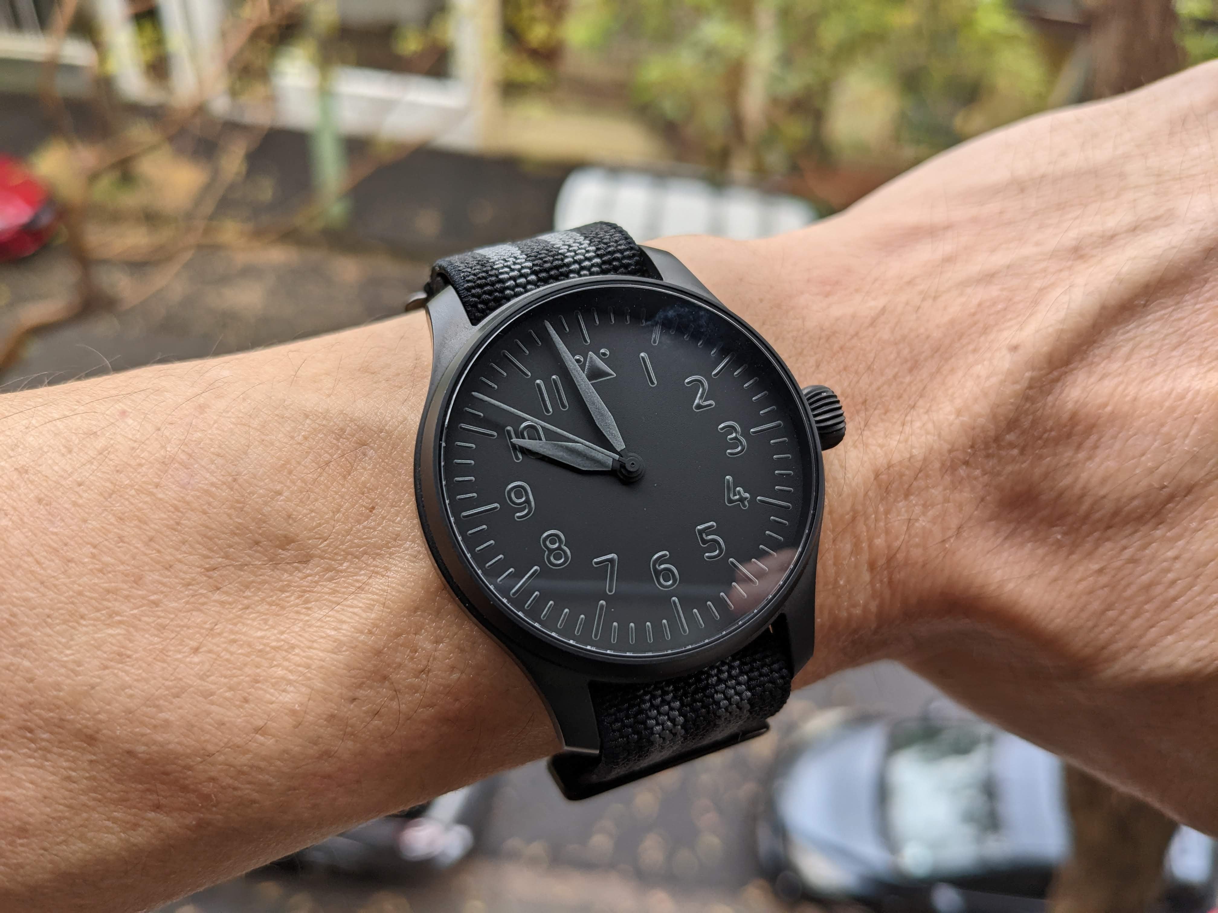 All-black minimalist watch with Arabic numerals and textile strap worn on wrist in outdoor garden setting